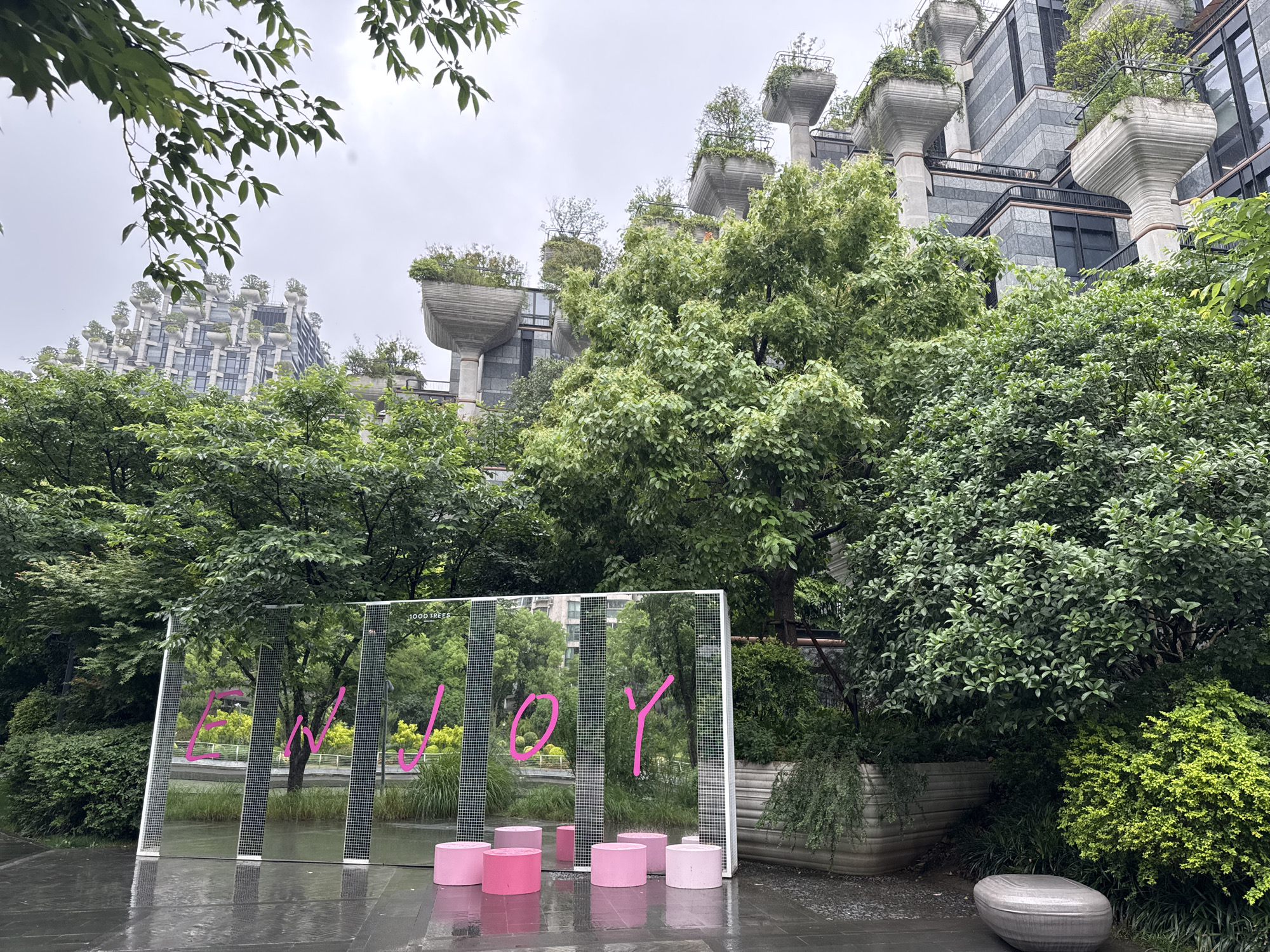 The image captures a scene in Putuo District, China, marked by the presence of the '1000 Trees' building, distinguished by its unique architectural design featuring numerous trees planted on its terraces and elevated pillars. In the foreground is a mirrored art installation displaying the word "JOY" in pink lettering, complemented by a series of pink cylindrical stools placed in front. The ground is wet, suggesting recent rain, and the surrounding environment is lush with greenery, including various trees and shrubs. A concrete bench sits off to the right. The weather appears overcast, casting a muted light over the scene.