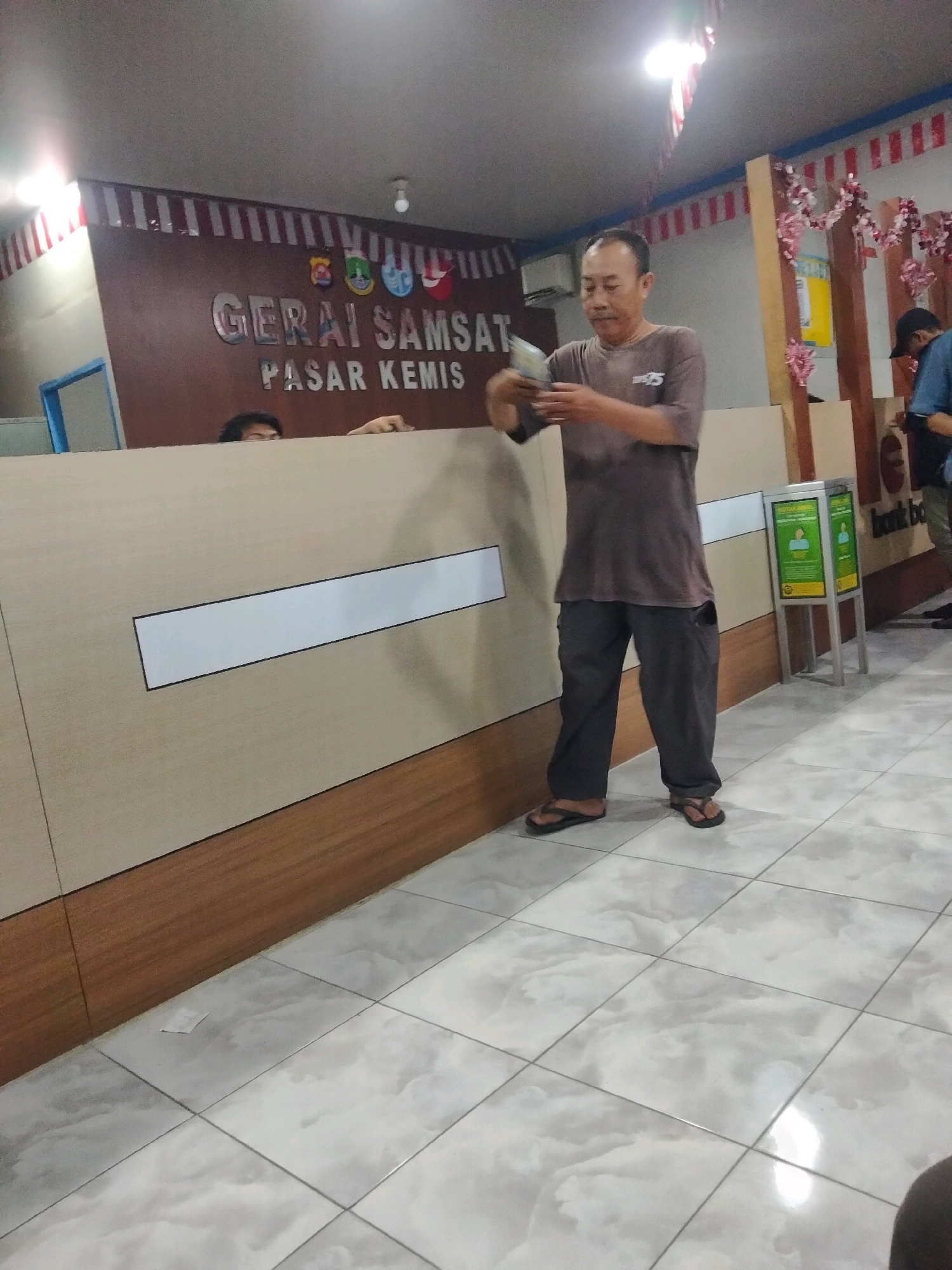 The image depicts an interior scene at a "GERAI SAMSAT PASAR KEMIS" location, which is a tax payment service outlet in Sukaasih, Indonesia. A middle-aged East Asian man, wearing a grey t-shirt and dark grey pants, stands near a counter, holding a stack of paper bills in his hands, appearing to count or examine them. He is wearing dark flip-flops. Behind him, partially visible behind the counter, are two individuals, one with dark hair peeking over the counter and another in a blue shirt to the right. The counter itself is a light beige color with a dark brown wooden base. A white rectangular panel with a black border is affixed to the counter. To the right of the man, a green and yellow information kiosk is visible. The floor is tiled with a marble-like pattern. The walls feature a brown backdrop with red and white striped decorations at the top. The overall lighting suggests an indoor environment.