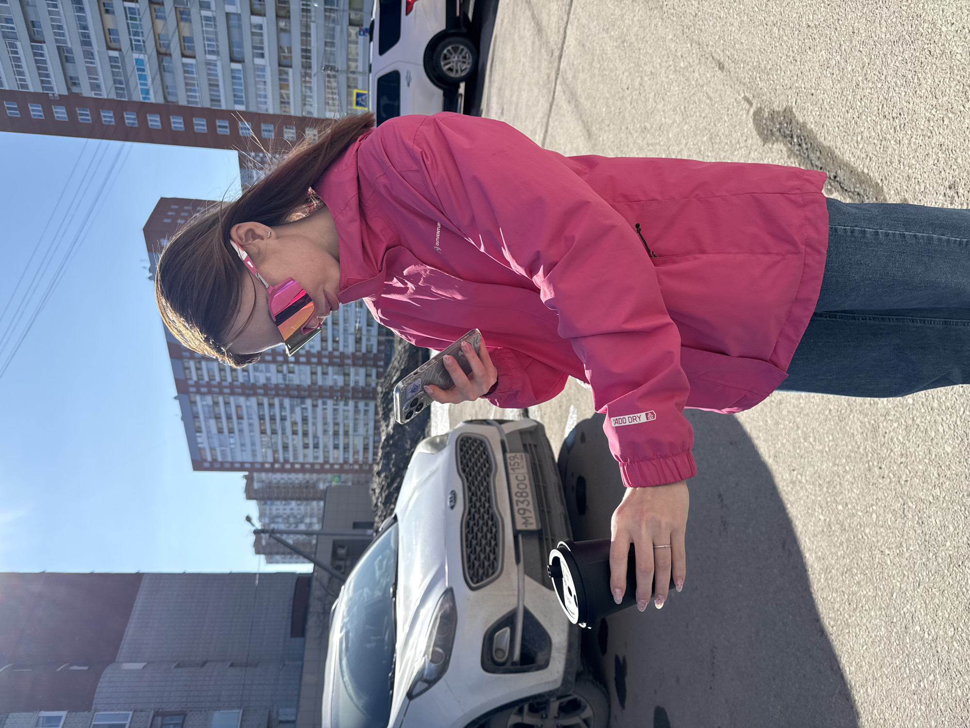 A woman in a pink jacket and jeans stands on a sunny street in what appears to be Berezniki, Russia, based on the car's license plate number. She holds a phone in one hand and a black travel mug in the other, seemingly looking at the mug. A white Kia car is visible in the background, with a building complex nearby. The sky is clear and blue.