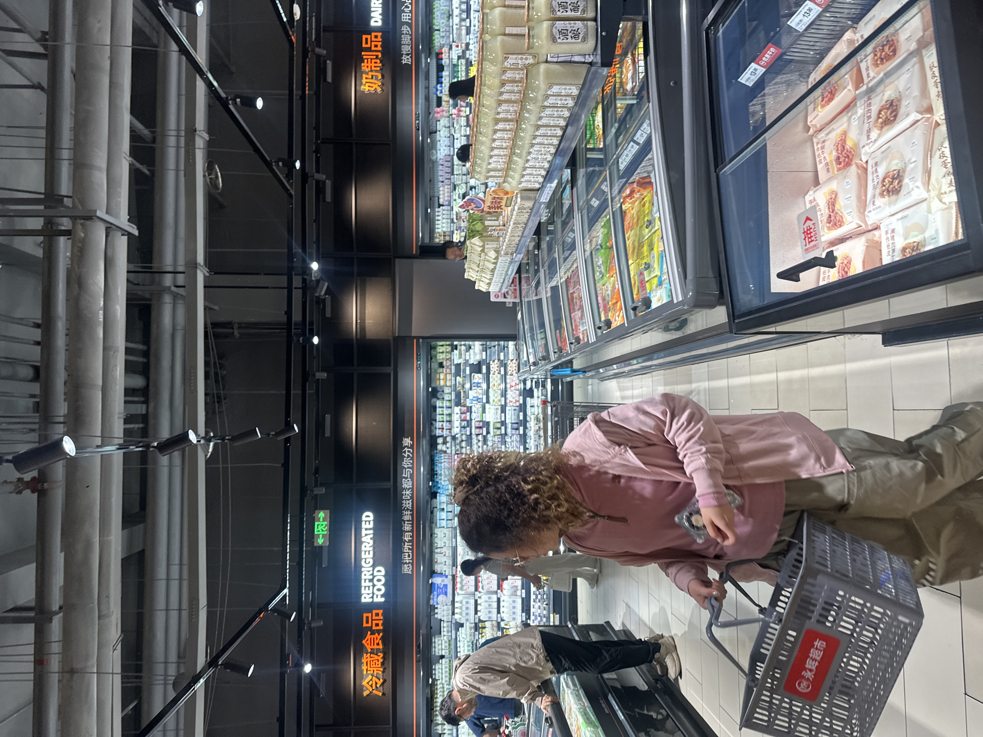 The image depicts a busy supermarket aisle in the Chongqing High-tech Zone, China, featuring a refrigerated section. In the foreground, a woman with curly brown hair, wearing a pink hooded jacket and tan pants, carries a grey shopping basket. She is facing away from the camera, looking towards the refrigerated displays. Behind her, a man in a beige jacket and dark pants is examining items in a display case. Further back, other shoppers are visible, as are shelves stocked with various food products, primarily dairy and refrigerated items, indicated by prominent signage in Chinese and English that reads "Refrigerated Food" and "Dairy." The overall environment is brightly lit, with overhead fluorescent lighting and spotlights illuminating the merchandise. The flooring appears to be tiled.
