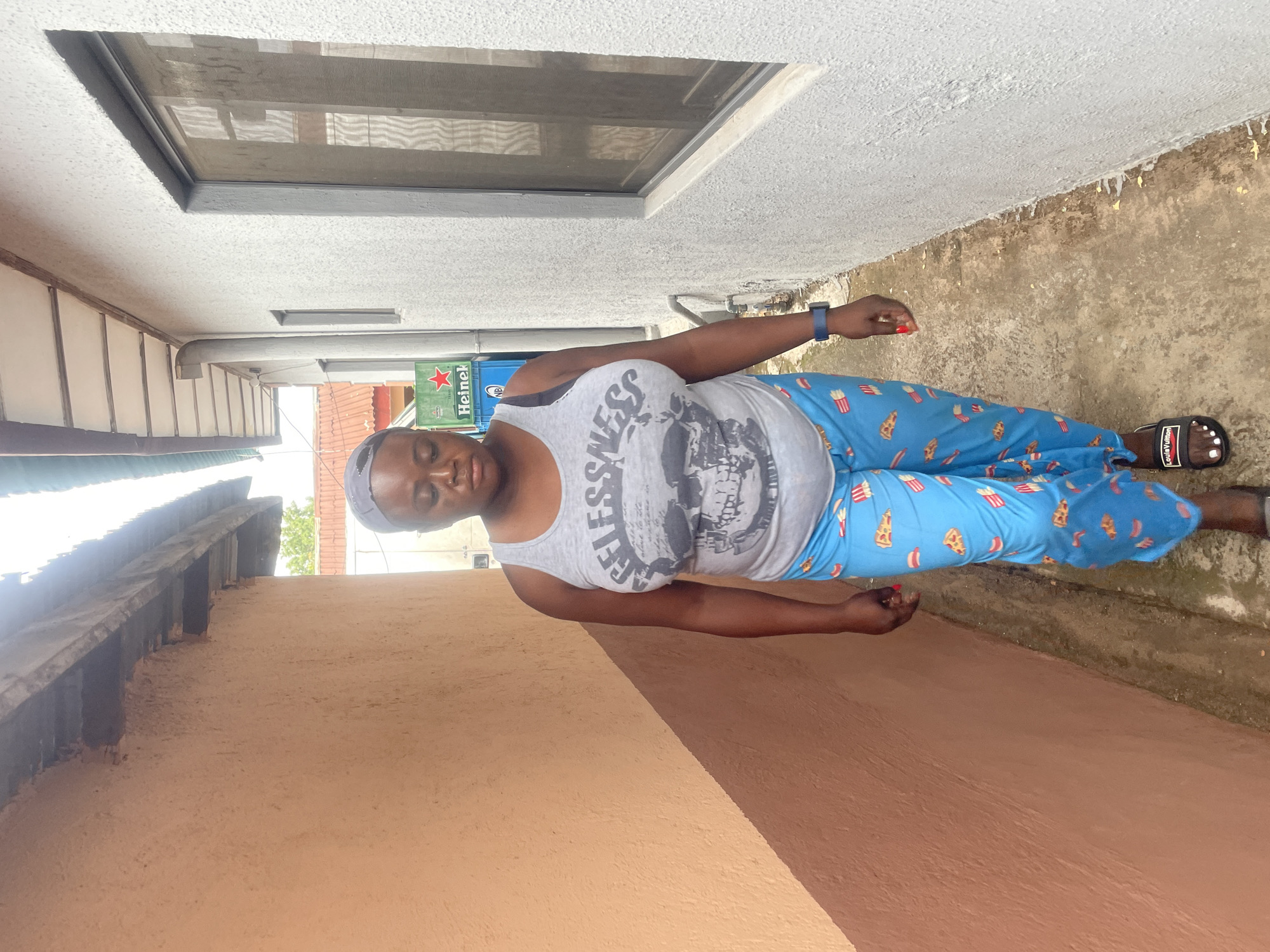 The image captures a female individual standing upright in a narrow outdoor passage or alleyway between two buildings in what appears to be a daytime setting. The person, with dark skin, is wearing a light grey tank top featuring the partially visible word "FELESSNESS" and a graphic of a skull. She pairs this with vibrant blue pajama pants patterned with images of pizza slices, hot dogs, and french fries. Black sandals with "Louis Vuitton" branding are on her feet, and a blue smart watch is visible on her left wrist. Her fingernails are painted red, and her head is covered by a grey cap or headwrap. Her expression is neutral and relaxed.
The immediate surroundings feature a light-colored, textured wall on the right, which appears to be painted concrete, leading up to a window with a mesh screen. Further along this wall, a series of green Heineken crates are stacked. To the person's left, a wall of a different reddish-brown or beige hue creates a contrasting backdrop. The ground surface is a mix of concrete and dirt, showing some discoloration and wear. Above the person's head on the left side, architectural elements suggestive of a building's underside or roof structure are visible.
The bright lighting suggests a clear, possibly sunny day. The scene conveys a casual, everyday moment in an urban or semi-urban environment, consistent with the provided location context of Jos, Nigeria.
