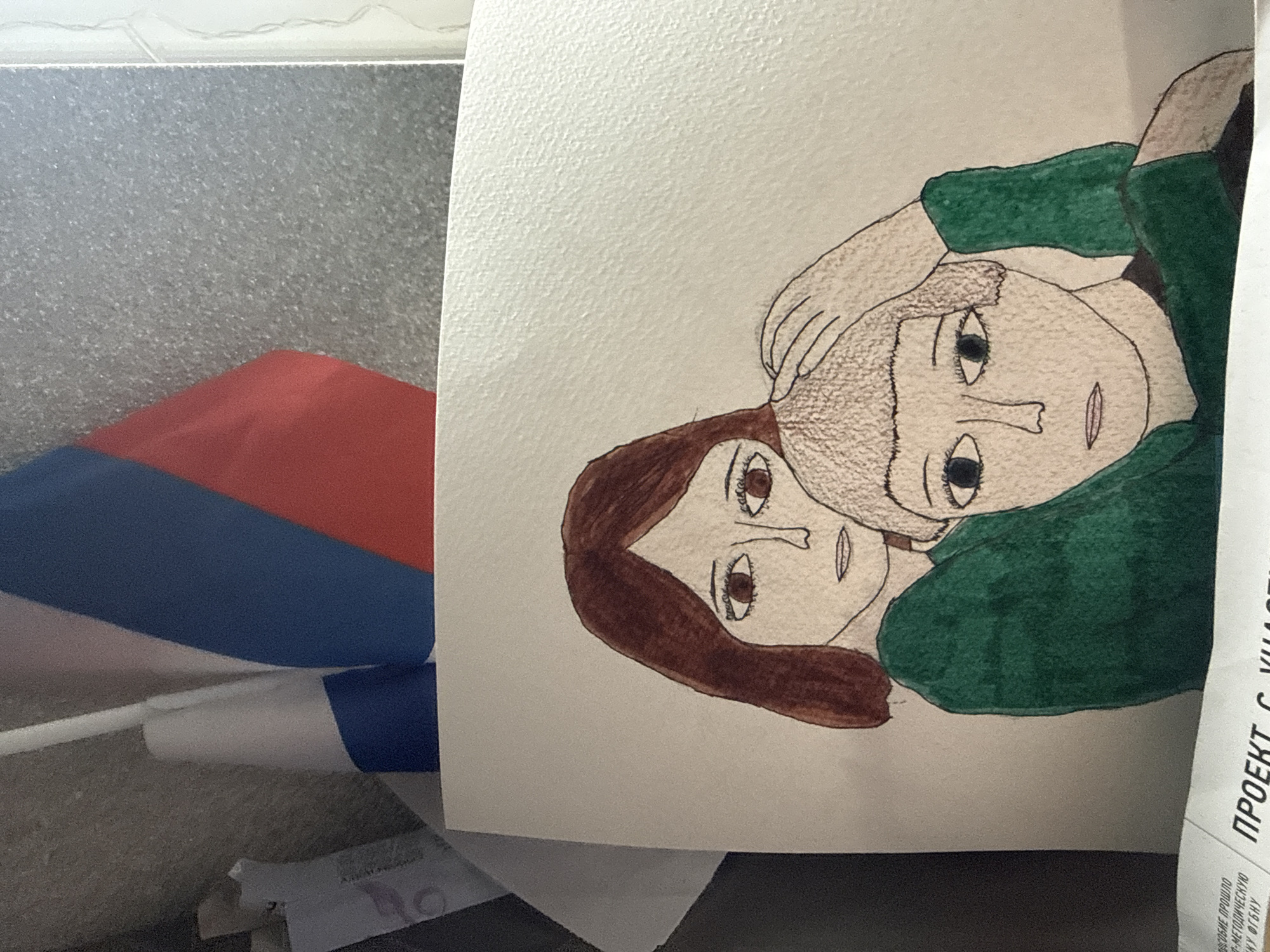 The image shows a drawing of two people embracing. One person has brown hair and the other has blonde hair. They are wearing a green shirt. The drawing is on white paper. In the background, there is a Russian flag and some crumpled paper. The location is unknown and cannot be determined from this image.