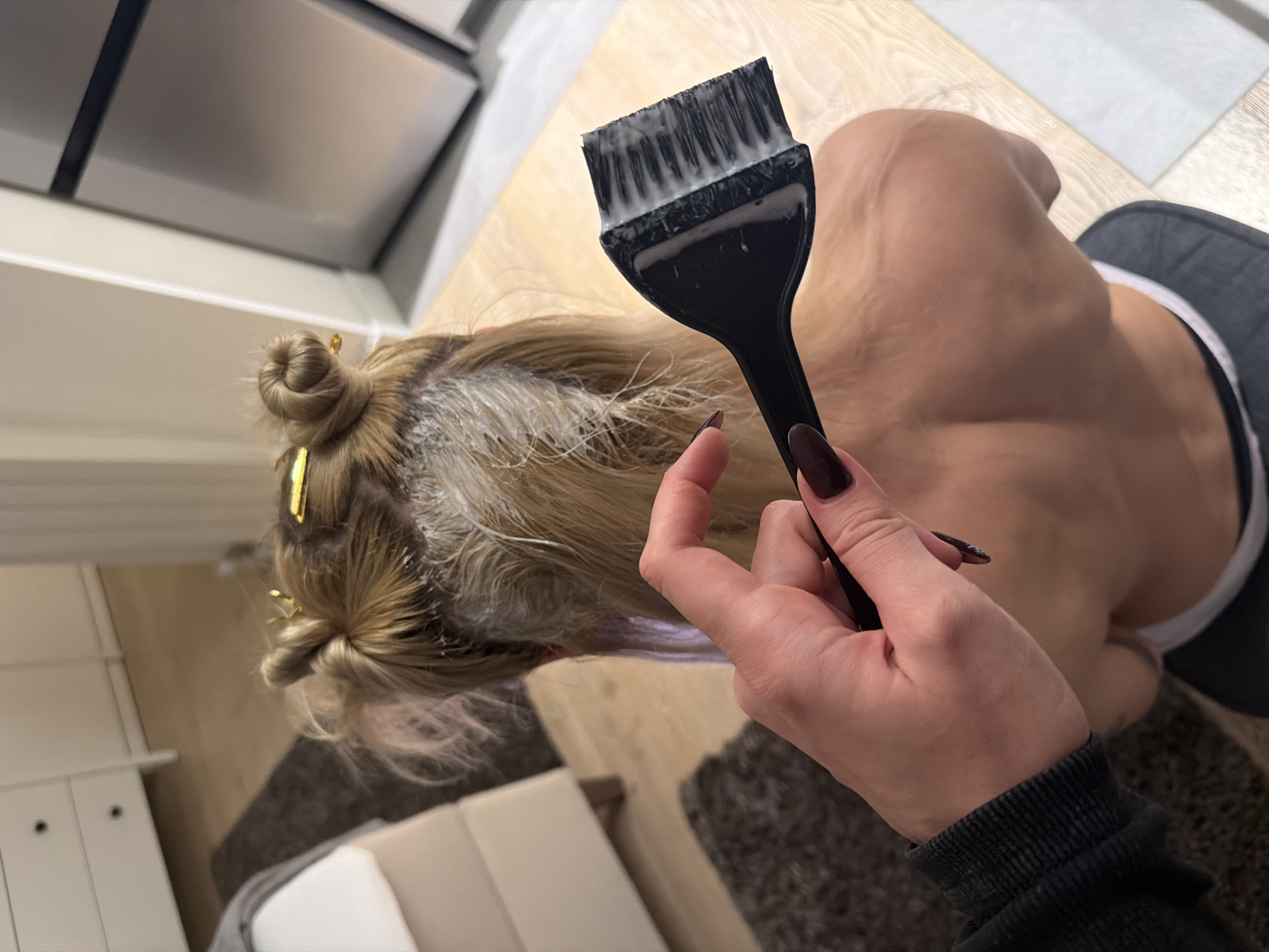 The image depicts an indoor residential scene in Belgrade, Serbia. The exact location within the city is indeterminate. A person with blonde hair is seated, viewed from behind, undergoing a hair treatment. Their hair is sectioned into two top buns secured with gold-colored clips, and a white, creamy substance is visible on the hair roots and scalp, consistent with hair dye or bleach application. The individual's bare shoulders and upper back are exposed, and dark-colored clothing with a white waistband is visible around their lower back. In the foreground, a hand with dark-polished fingernails is holding a black hair dye brush, loaded with the same white, creamy substance, poised for application. The background features light wooden flooring, a dark shaggy rug, a light-colored sofa, a white dresser with two black-knobbed drawers, and a partial view of a stainless steel appliance, likely a refrigerator or oven.