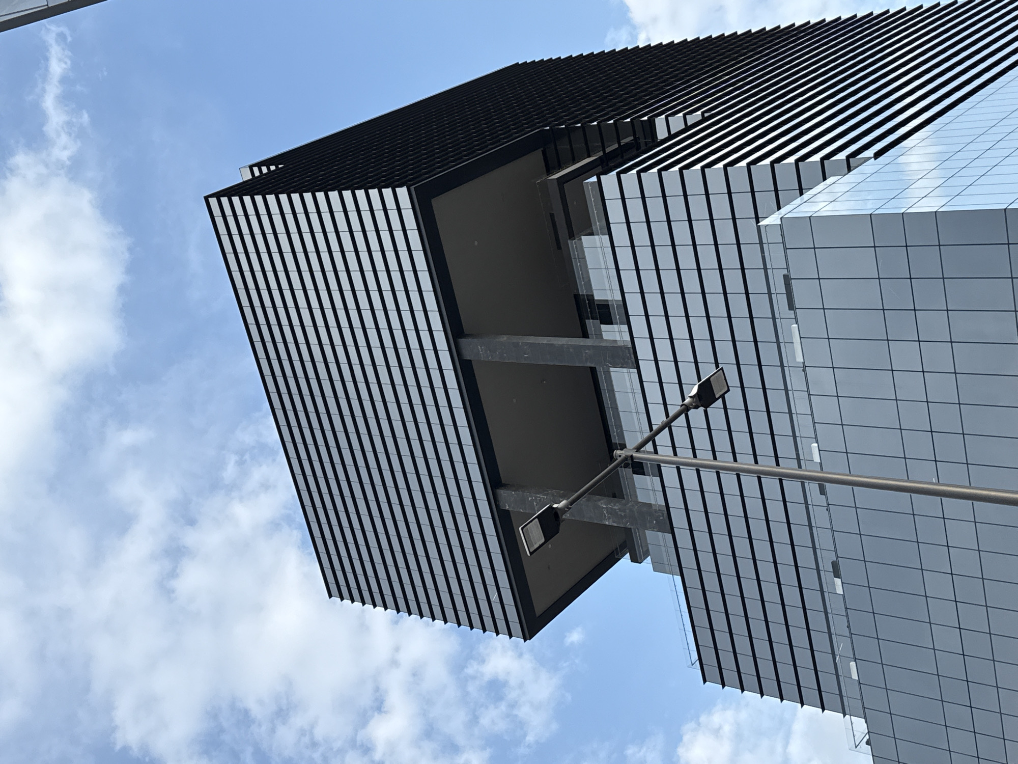 The photograph presents a low-angle view of a modern building with a distinctive architectural style. The building features a facade composed of horizontal black and white stripes, creating a striking visual pattern. A section of the building has a cantilevered design, extending outwards with visible support structures underneath. Adjacent to the striped section, another part of the building is clad in a grid of reflective, square panels.
A street lamp is positioned near the building, intersecting the view of the facade. The sky is partially visible in the background, exhibiting a blue hue with scattered white clouds.
The location is Puebla, Mexico.