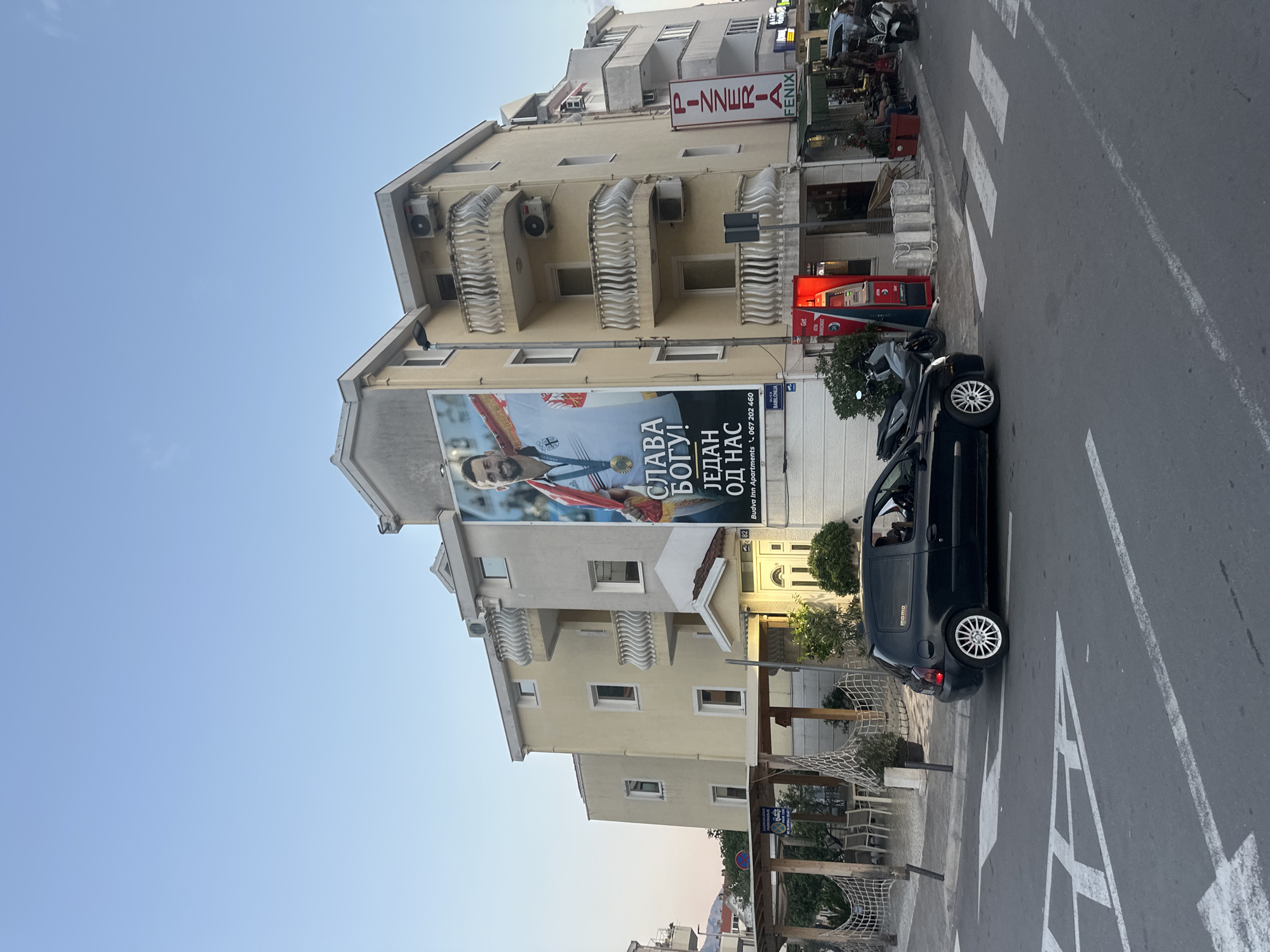 Here's a detailed description of the image:
**Setting:** The image is taken in Budva, Montenegro. The scene depicts an urban setting, with buildings lining a street. The sky is clear and blue.
**Buildings:** A multi-story building dominates the scene. It has a cream or light yellow facade, with decorative white railings on the balconies. A large billboard is mounted on the building, displaying a picture of a man wearing a Serbian sports uniform and holding a medal. Text on the billboard reads "SLAVA BOGU! JEDAN OD NAS" along with "Budva Inn Apartments". Above the billboard and closer to the street, a sign for "PIZZERIA FENIX" is visible. There's also an ATM from Babilonija nearby.
**Street Scene:** A black car is parked on the side of the road. Crosswalk markings are visible on the asphalt.
**People:** The primary human subject is on the billboard. In the background some persons can be seen next to Pizzeria Feniks.
**Objects:** Key objects include the billboard, the car, the pizzeria sign, the ATM, and architectural elements like balconies and windows. There is mesh netting visible near a pedestrian area.
**Overall Impression:** The image shows a typical street scene in Budva, blending residential/commercial buildings with everyday traffic and amenities.