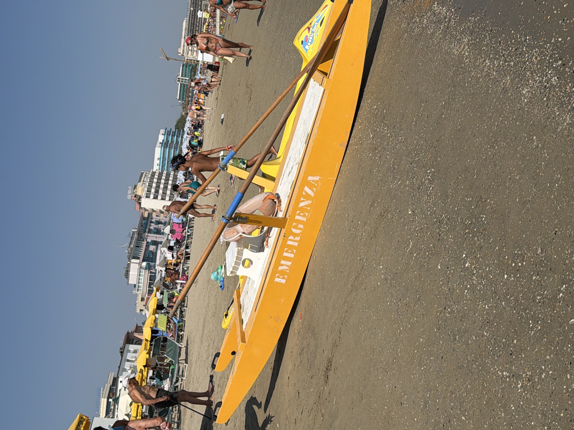 An outdoor, eye-level, wide shot captures a bright, sunny day on the beach in Gatteo, Italy. The focal point is a bright orange, life-saving boat positioned on the sand in the foreground, angled towards the viewer's left. The word "EMERGENZA" is prominently displayed in white lettering on the side of the boat. Two wooden oars with blue accents are resting across the boat.
The background reveals a crowded beach with numerous people scattered across the sand, many of whom are engaged in activities like sunbathing or walking. Beach umbrellas, lounge chairs, and buildings line the coast, indicating a developed tourist area. The sand itself appears dry and coarse, with scattered pebbles and shells. The sky is a clear, bright blue.