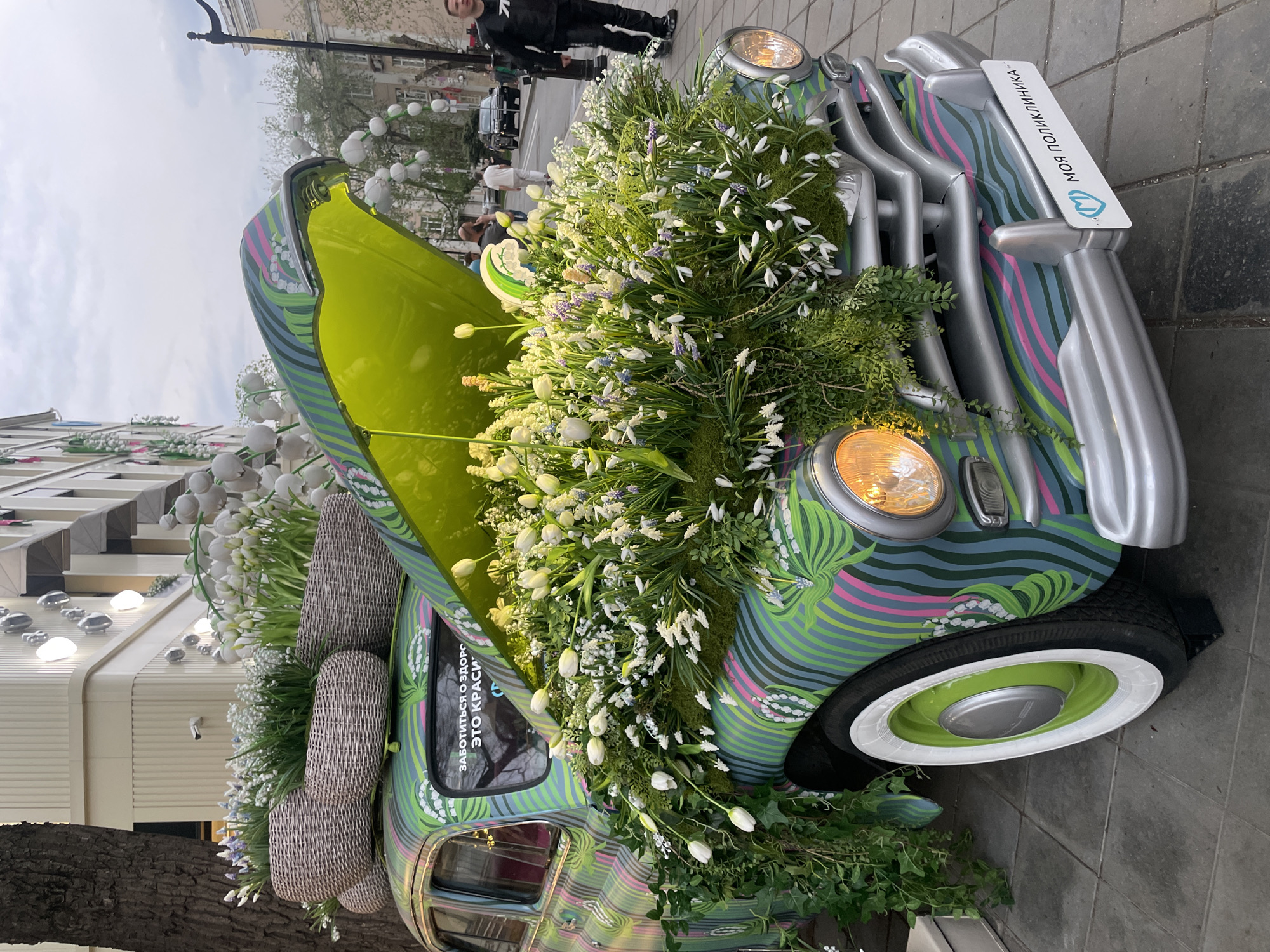 A highly decorated car is on display on a Moscow, Russia, street. The car, which has a lime green hood and is covered in green, blue, pink, and gray stripes, is adorned with white flowers. Additional white flowers are on top of two woven baskets located at the rear of the car. The car's front grill is silver. The license plate reads, "МОЯ ПОЛИКЛИНИКА." The car's headlights are on. A person in a black jacket and black pants is walking on the sidewalk behind the car. Buildings can be seen in the background, and a few people are standing on the sidewalk. Round, white lights are hanging on the buildings on the opposite side of the street.