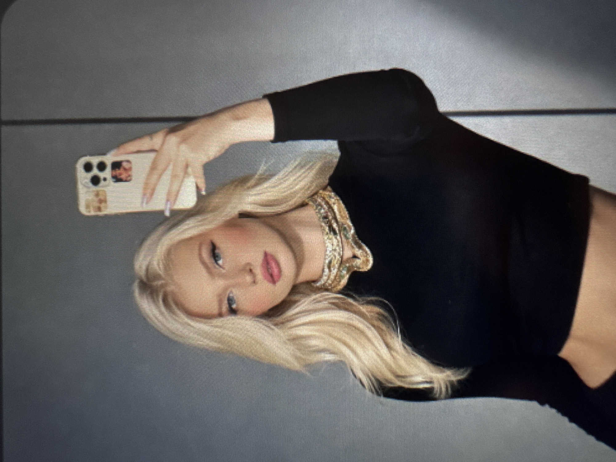 A young woman with long, straight blonde hair is featured in a posed portrait, likely a selfie or mirror shot, against a plain, dark gray background. Her head is tilted slightly, and she looks directly towards the camera with a neutral expression, enhanced by visible eye makeup and pink lipstick. She wears a black long-sleeved crop top and a prominent, ornate gold-colored necklace with an intricate, possibly layered design.
She holds a light-colored smartphone, consistent with an iPhone model due to its camera array, with her right hand. The phone's case appears decorative, featuring small, indistinct images or stickers on the back. Her fingernails are painted a light color. The setting appears to be indoors, possibly against a studio wall or a simple room background, with no distinguishing location cues. There is no visible text in the image.
