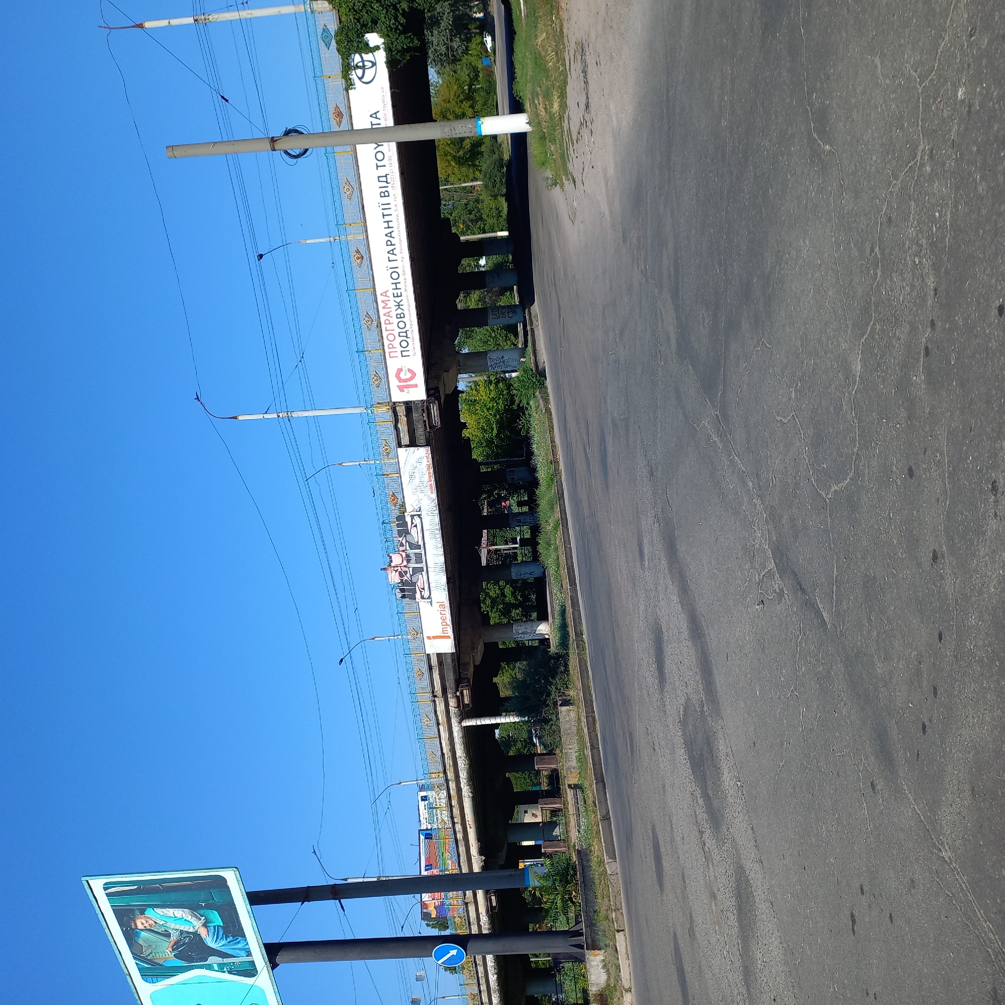 This image shows an overpass on a sunny day in Kropyvnytskyi, Ukraine. The overpass is supported by concrete pillars and has a blue and yellow decorative railing. Various advertisements are displayed on the side of the overpass, including one for Toyota and another with a picture of a man in a car. Below the overpass, there is a road with some traffic signs, including a blue sign with a white arrow indicating a turn. There are also trees and vegetation growing around the base of the overpass. The road surface appears to be asphalt and shows signs of wear and tear. The sky is clear and blue. There are no people clearly visible in the image.