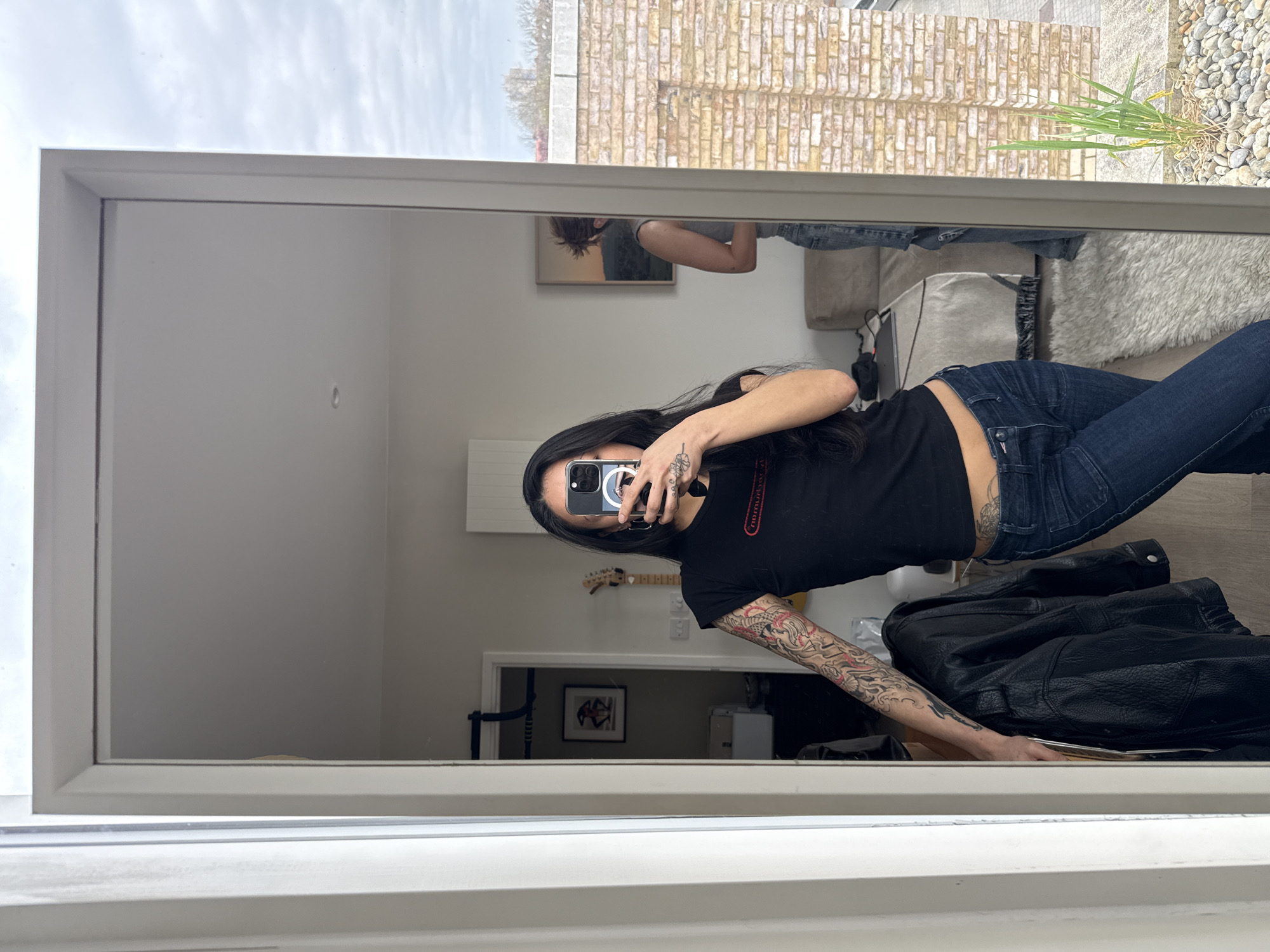 The image is a mirror selfie of a person with long dark hair, wearing a black t-shirt and blue jeans, with tattoos on their arm. The reflection shows a room interior with light-colored walls, a guitar, a framed picture on the wall, and a door leading to another room. A black leather jacket is placed on a surface in the foreground. The mirror also reflects an exterior view with a brick building and a pebbled ground area with plants. The image does not provide enough information to determine the specific location in London, United Kingdom.