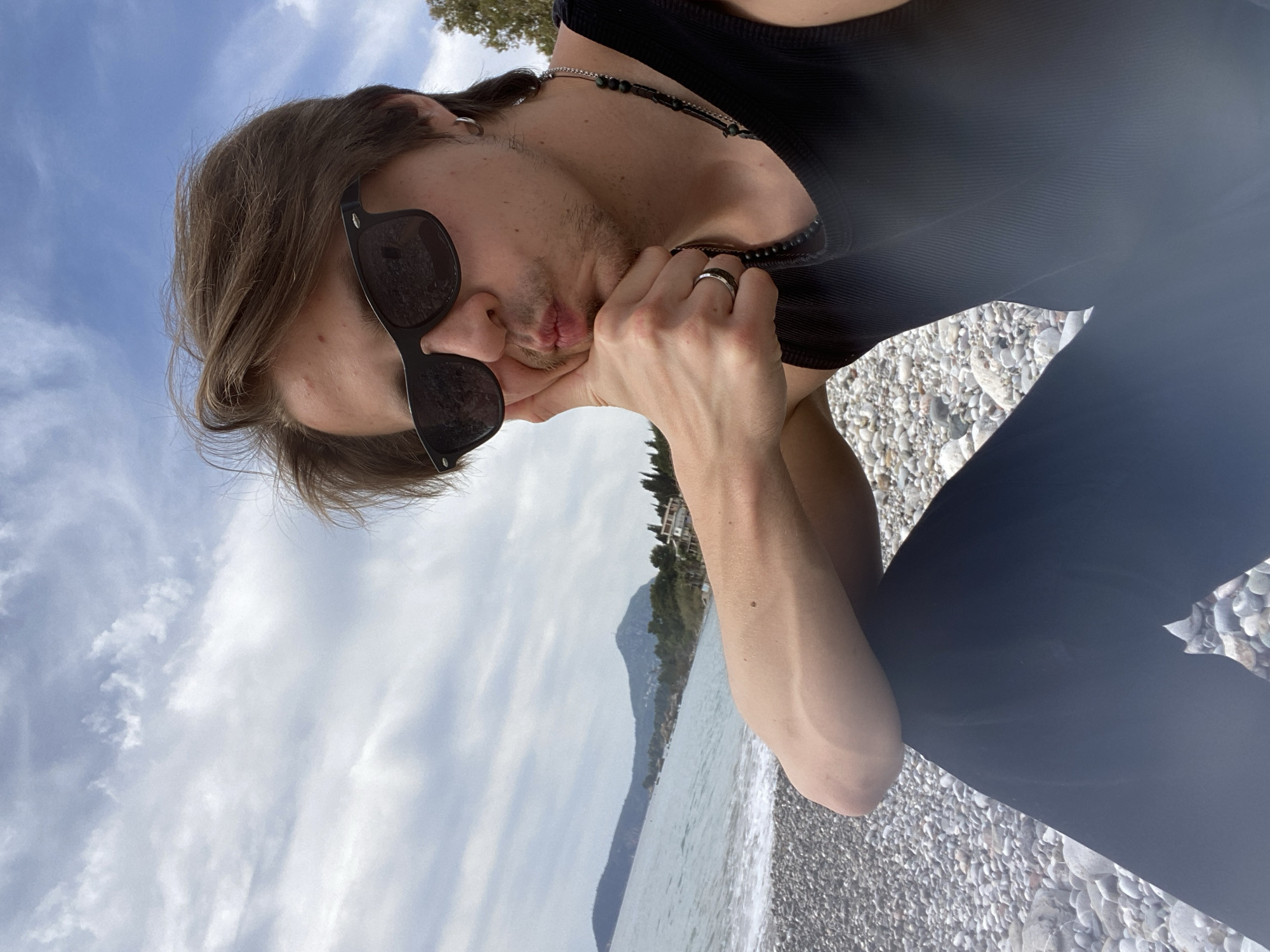 A young, light-skinned male with brown hair and sunglasses is on a pebble beach in Bar, Montenegro. He's wearing a black tank top, black pants, and a beaded necklace. His face rests on his hand, displaying a silver ring on his finger. Behind him is the Adriatic Sea, a distant mountain, and cloudy sky. The beach is made of small, light-colored pebbles. The scene is captured from a low angle, looking upwards.