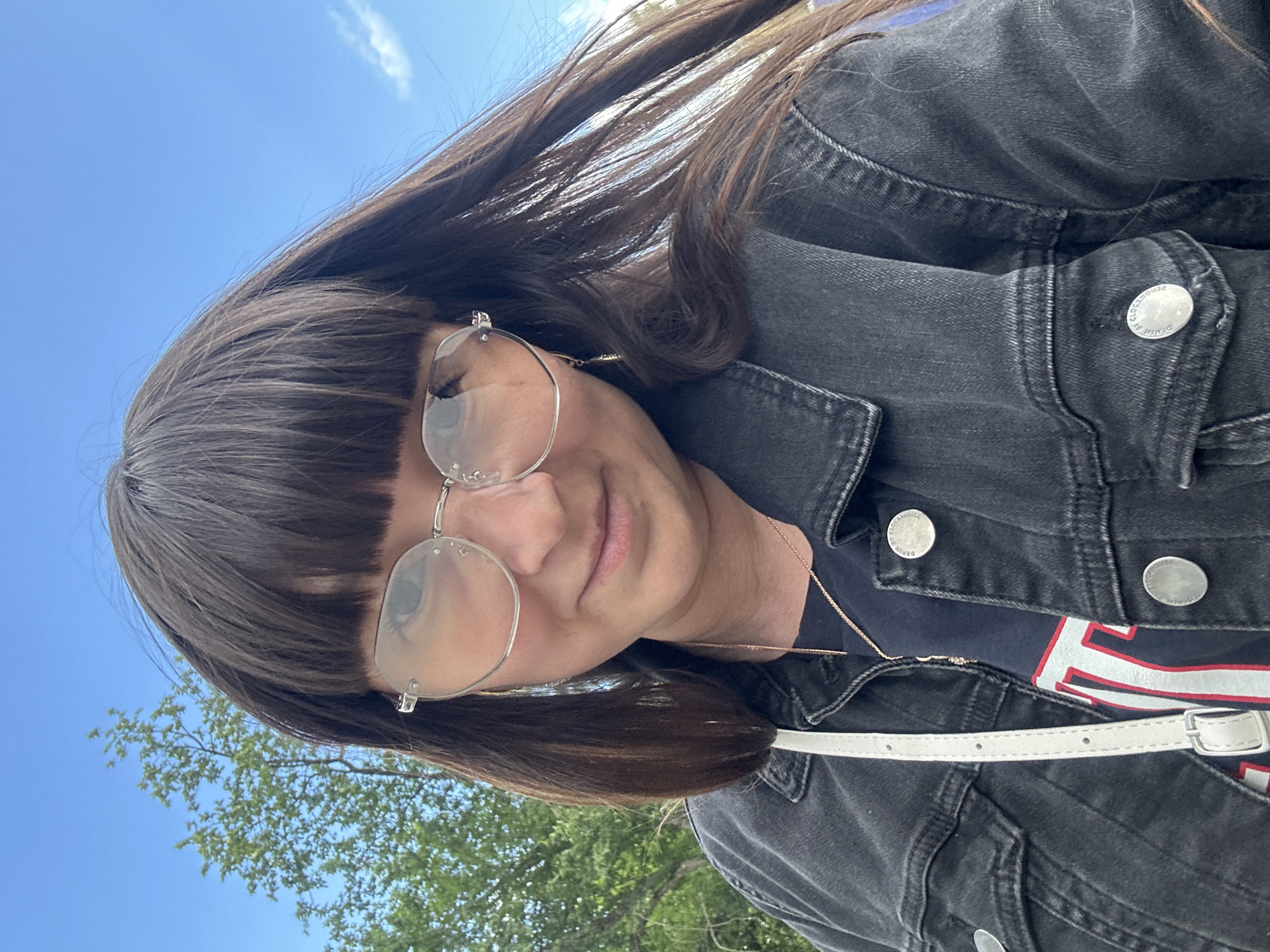 A selfie shows a young woman with long, dark brown hair and bangs, wearing large, round, silver-rimmed glasses. She is also wearing a dark denim jacket over a dark t-shirt with red and white lettering. A white purse strap is visible across her chest. The background is a bright blue sky with a few wispy clouds and the top of a green tree. The image appears to have been taken outdoors on a sunny day. The location is Chaikovsky, Russia.