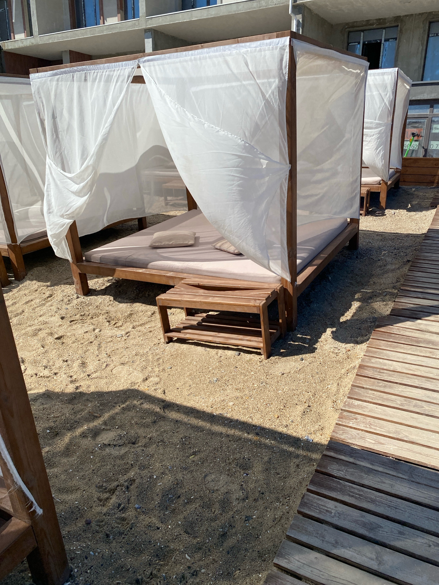 This is a medium shot, eye-level photograph taken outdoors on a sunny day, showing a row of beach cabanas in Odesa, Ukraine. The cabanas are made of wood with white sheer curtains that hang from the top. Each cabana has a cushioned bed and a small wooden bench or table next to it. The cabanas are situated on a sandy beach, with a wooden walkway running alongside them. In the background, a multi-story building with windows is visible. The scene appears to be a resort or a beach club. There are no people in the image, and no action is taking place. The lighting suggests it is daytime, with strong sunlight casting shadows.
