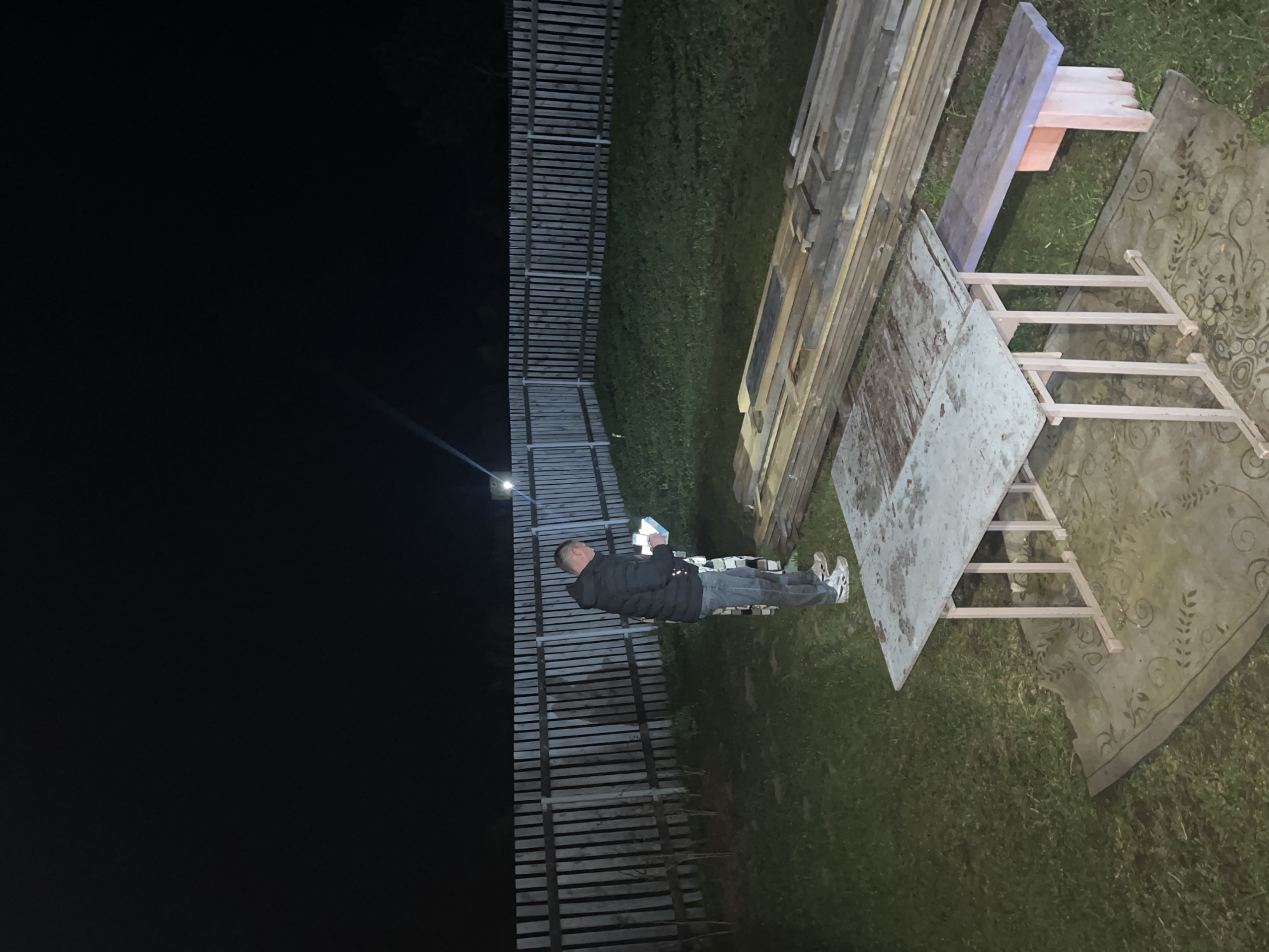 The image depicts a man standing in a yard in Гаревая, Russia, next to a wooden fence and various objects. The man is wearing a dark jacket and jeans. He appears to be holding something small in his hands, possibly a mobile phone. Next to him are stacks of wooden boards and a metal table with a light-colored frame standing on top of a patterned mat on the green grass. The left side of the image is dark, suggesting it's nighttime or a dimly lit area. A single light source is visible near the fence.