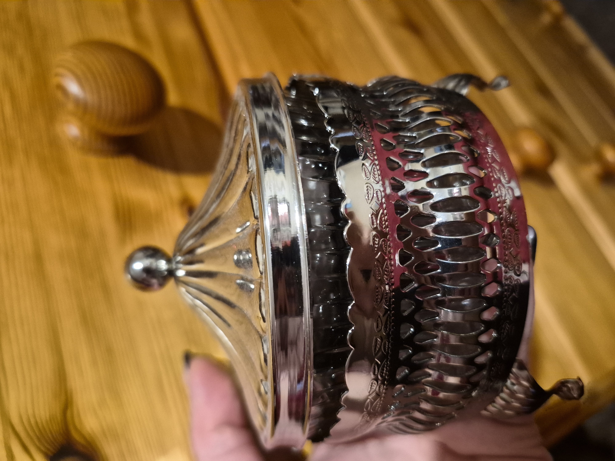 A silver decorative container with a lid is held in a person's left hand against a backdrop of wooden planks. The container has intricate metalwork, including a fluted lid topped with a small sphere, a decorative band around the rim, and an openwork design on its body with small feet. The wooden planks are arranged horizontally and have a warm, light brown color. The overall scene is well-lit, with some shadows visible on the wooden surface. The image appears to be a close-up shot of the decorative object. The location of Sandwell, United Kingdom is not discernible from the image.