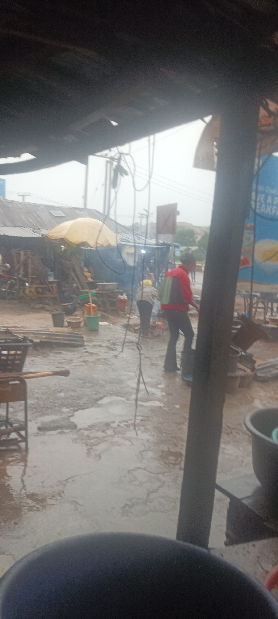 The image depicts an outdoor scene in Kuru Dungus, Nigeria, characterized by wet conditions, likely after rainfall. In the foreground, the bottom of a dark-colored bucket is visible, along with a metal pole structure. The middle ground reveals several people engaged in activities amidst puddles of water. A woman wearing a yellow head covering and dark pants appears to be tending a fire, while a person in a red jacket and dark pants stands nearby. Further back, there's a structure covered with a yellow umbrella, possibly a makeshift stall or cooking area. The backdrop shows power lines and indistinct buildings in the distance, suggesting a semi-urban environment. A sign is partly visible on the right side, offering breakfast.