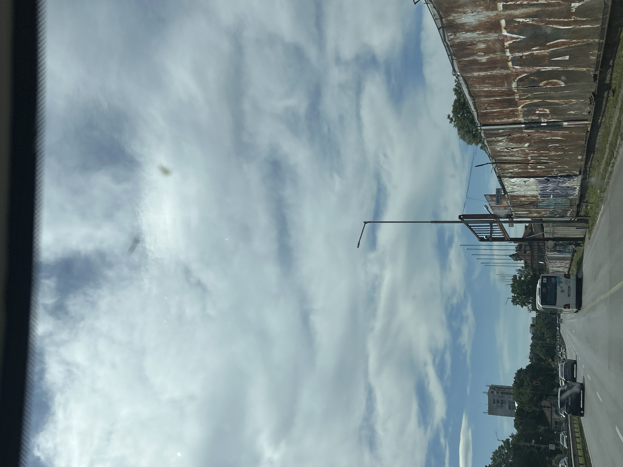 The image shows a street scene in Sofia, Bulgaria, taken from inside a vehicle. The sky is partly cloudy. Along the right side of the road, there is a rusted and graffiti-covered wall, a street pole, some buildings, trees, and moving vehicles, including a white bus and several cars. The asphalt road has yellow lines. The scene is captured at eye-level. The interior of the vehicle is visible on the left edge of the frame.