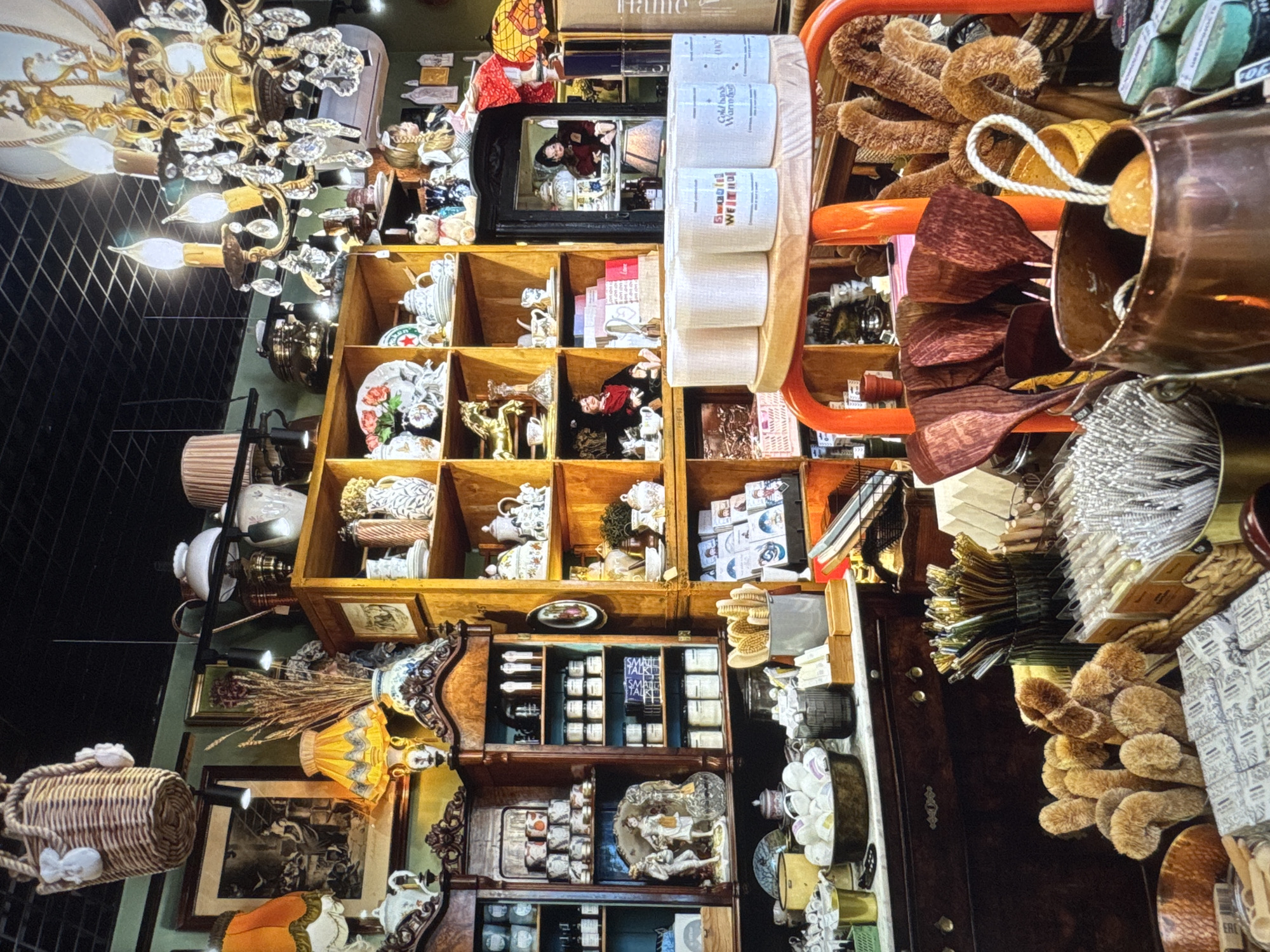 The image depicts an indoor scene, likely a shop, filled with various decorative and functional items. In the foreground, there are several objects including a copper pot, wooden spatulas, brushes, and packaged goods. Behind these, a dark wooden cabinet is visible with drawers and shelves displaying more items.
Further back, wooden shelving units are packed with an assortment of ceramics, teapots, vases, and decorative figures. Some shelves also hold baskets and framed pictures. A significant feature is a large, ornate crystal chandelier hanging from the ceiling. Several lamps with fabric shades are also present throughout the space. The overall impression is of a cluttered yet curated collection of vintage or antique items, possibly within a boutique or antique store. The backdrop consists of green walls and a dark, tiled ceiling. There are no people clearly visible in the image. The exact location in Novinki, Russia, is not discernible from the visual information provided.