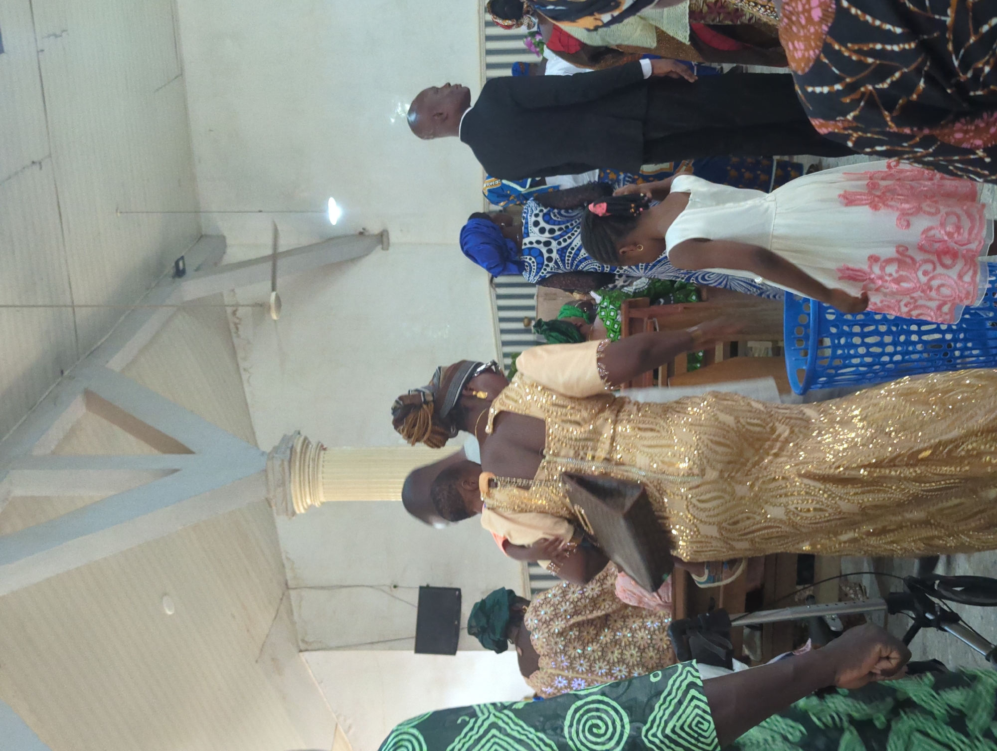 The image shows a group of people gathered indoors, possibly in a church or community hall. The environment appears to be a bit dimly lit with white walls and a light-colored ceiling with exposed structural beams. In the foreground, a woman in a gold sequined dress is holding a child. She is carrying a dark brown handbag. To her left, another person is visible in a green patterned garment. To the right of the woman and child, a young girl in a white dress with pink floral accents is standing near a blue plastic basket. Further in the background, several other individuals are seen, some in traditional African attire, including a man in a black suit. A ceiling fan and lights are visible, suggesting an indoor setting. The overall impression is of a social gathering or religious event.
