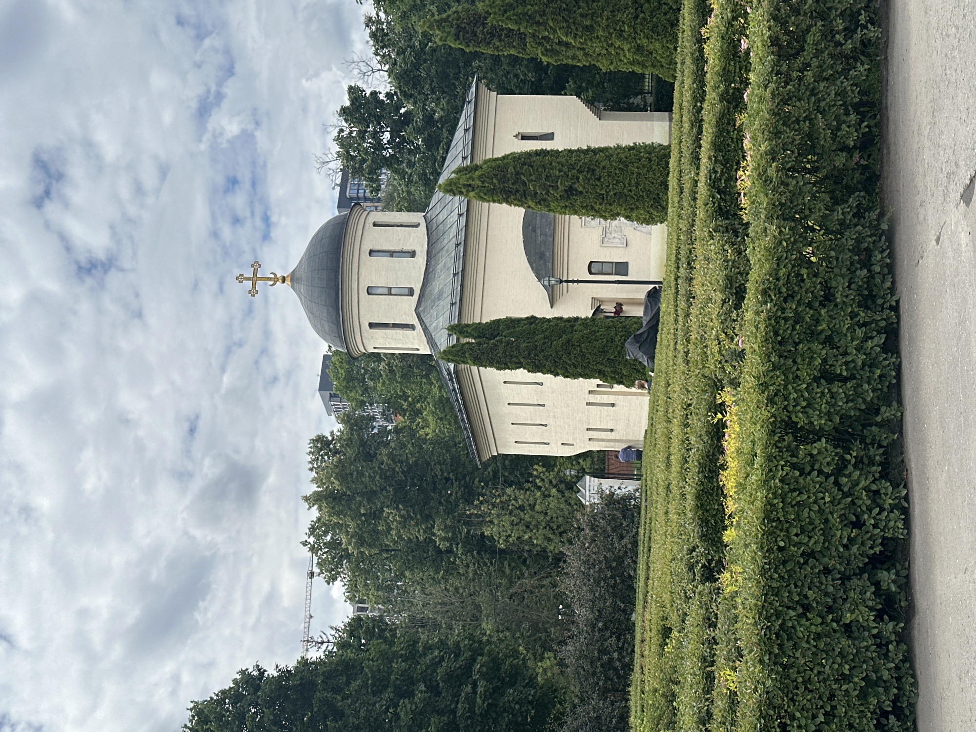 An exterior, medium shot captures a light-colored church with a dark dome and a gold cross against a cloudy sky. The church is surrounded by lush green trees and manicured hedges arranged in rows. In the foreground, a black tarp is draped over an unseen object on the ground. Two individuals, one wearing a striped shirt and the other with darker clothing, are visible near the church. A construction crane is visible in the distance amongst the trees. The scene appears to be a public park or garden.