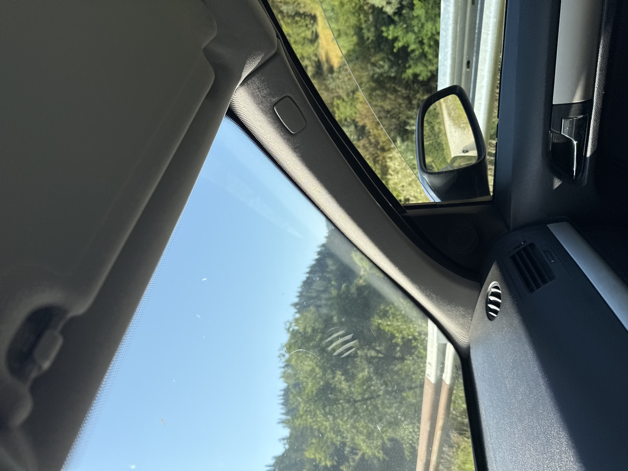 The image shows the interior of a car looking out the windshield and side window. The car is on a road, with trees and hills visible outside. The sky is clear and blue. The dashboard and side panel of the car are visible. There is a side view mirror showing a road and trees. This scene is likely from a rural area, possibly near Madan, Bulgaria, given the mountainous terrain and vegetation. There are no people or specific objects that place it precisely within the city of Madan, but the environment is consistent with its surrounding natural landscape.