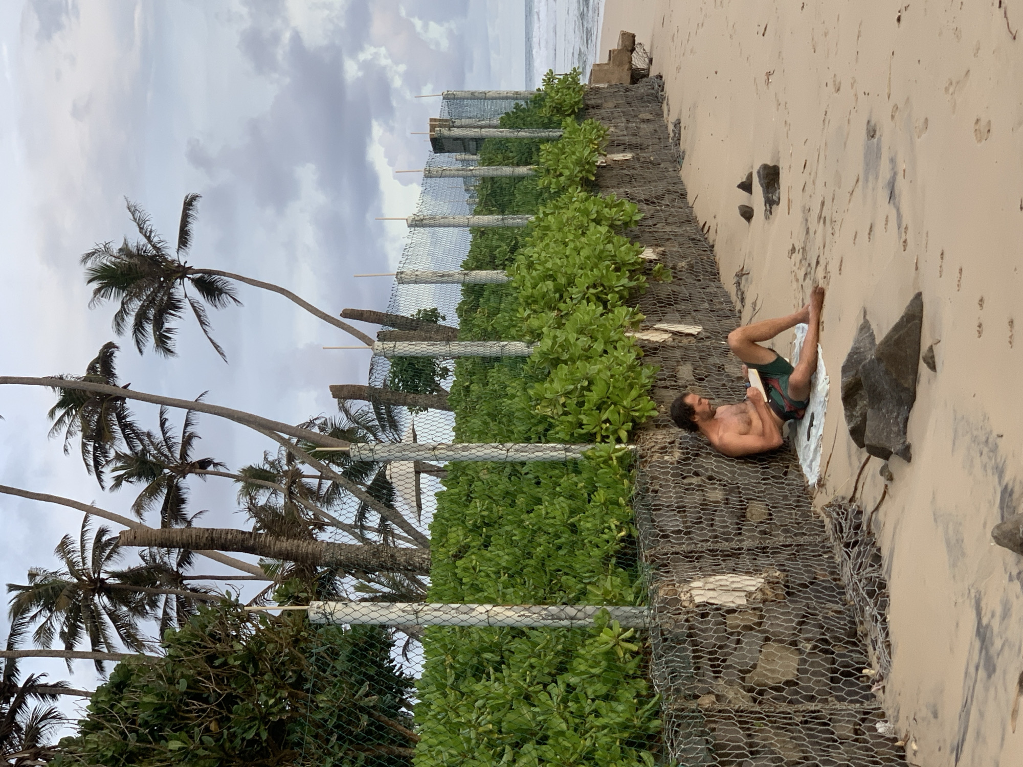 The image depicts a man relaxing on a sandy beach in Kathaluwa, Sri Lanka, under an overcast sky. The man is lying on his right side, propped up on his elbow, and appears to be reading a book or looking at a small device. He is shirtless, with a beard, and wears colorful swimming trunks.
Behind the man, running across the mid-ground, is a long coastal protection structure, likely a gabion wall made of wire mesh filled with rocks, which is partially covered with lush green vegetation. Numerous tall palm trees with dark fronds rise from behind this barrier, silhouetted against the cloudy sky. To the man's right, the sandy beach stretches towards the ocean, where gentle waves can be seen in the distance. Scattered dark rocks are visible on the sand near the man. The sky is largely cloudy, suggesting an overcast day or possibly late afternoon. The scene conveys a sense of calm and relaxation. No visible text is present.
