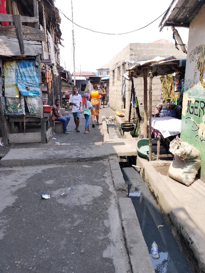 The image depicts a narrow, unpaved street or alleyway in Oworonshoki, Nigeria, under clear skies. The ground consists of uneven, dark-colored concrete and dirt, with scattered refuse, including a white wrapper. An open concrete drainage ditch runs along the right side of the street, containing dark, murky water and a clear plastic bottle.
On the left side, a weathered wooden structure, potentially a stall or building facade, stands. Large, discolored woven sacks, one blue and one white with text, are hung on its front. A male individual, dressed in a red sleeveless shirt and blue jeans, is seated on a low wooden stool next to this structure. A red plastic basin is visible nearby. Further down the left, other informal structures and what appears to be merchandise are present.
On the right side, a partially constructed concrete block building is visible, alongside a makeshift market stall supported by wooden posts and covered by a corrugated metal sheet. Colorful fabrics and other goods are displayed within this stall. A table covered with a white cloth holds additional merchandise. A green plastic basin rests on the ground near the stall, and a light-colored burlap-like sack sits on a concrete ledge bordering the drainage ditch. A wall painted with a yellow tiger and the word "TIGER" in green is also visible on the right.
Four individuals are walking in the middle ground, away from the foreground. The closest, a male, wears a white t-shirt and light shorts, holding a white object. Behind him, a female wearing a light-colored top and dark pants has her hair covered by a pink scarf. Further back, another female, dressed in a bright orange top and a patterned skirt, carries a large, dark object on her head. A fifth, less distinct figure is visible further down the street. Overhead, power lines traverse the scene. The structures on both sides are varied, including wooden planks, corrugated metal, and concrete blocks, reflecting an informal urban environment.