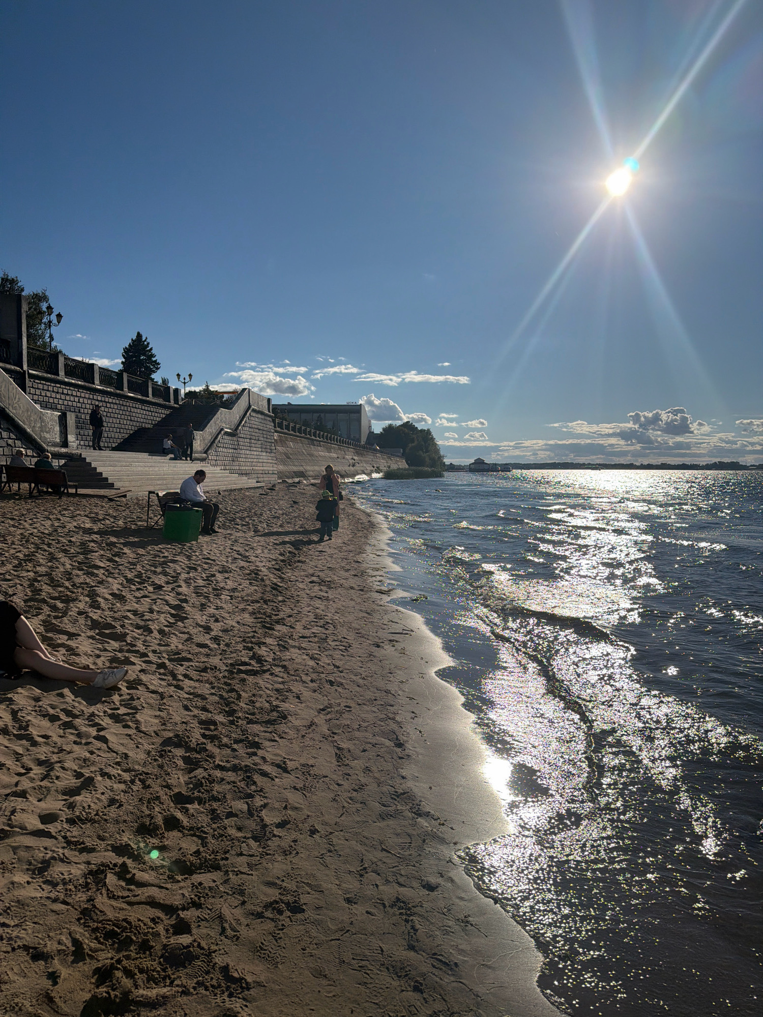 The image shows a sandy beach on a sunny day in Samara, Russia, likely near the Volga River embankment. In the foreground, sand is textured with footprints. To the left, a person's legs, wearing white sneakers and denim shorts, are visible, with their body out of frame. Further back, a man in a white shirt sits on a green bench, looking down. Beside him, a person in a dark shirt sits on another bench, and a small child in a green hat is walking towards the water. A woman stands near the child, facing the water. Behind them, a paved embankment with stairs and railings rises, with several people visible on the stairs. Buildings and trees are situated along the top of the embankment. The sun shines brightly in the upper right corner of the image, casting lens flares and creating a sparkling effect on the rippling water. The sky is blue with scattered white clouds.