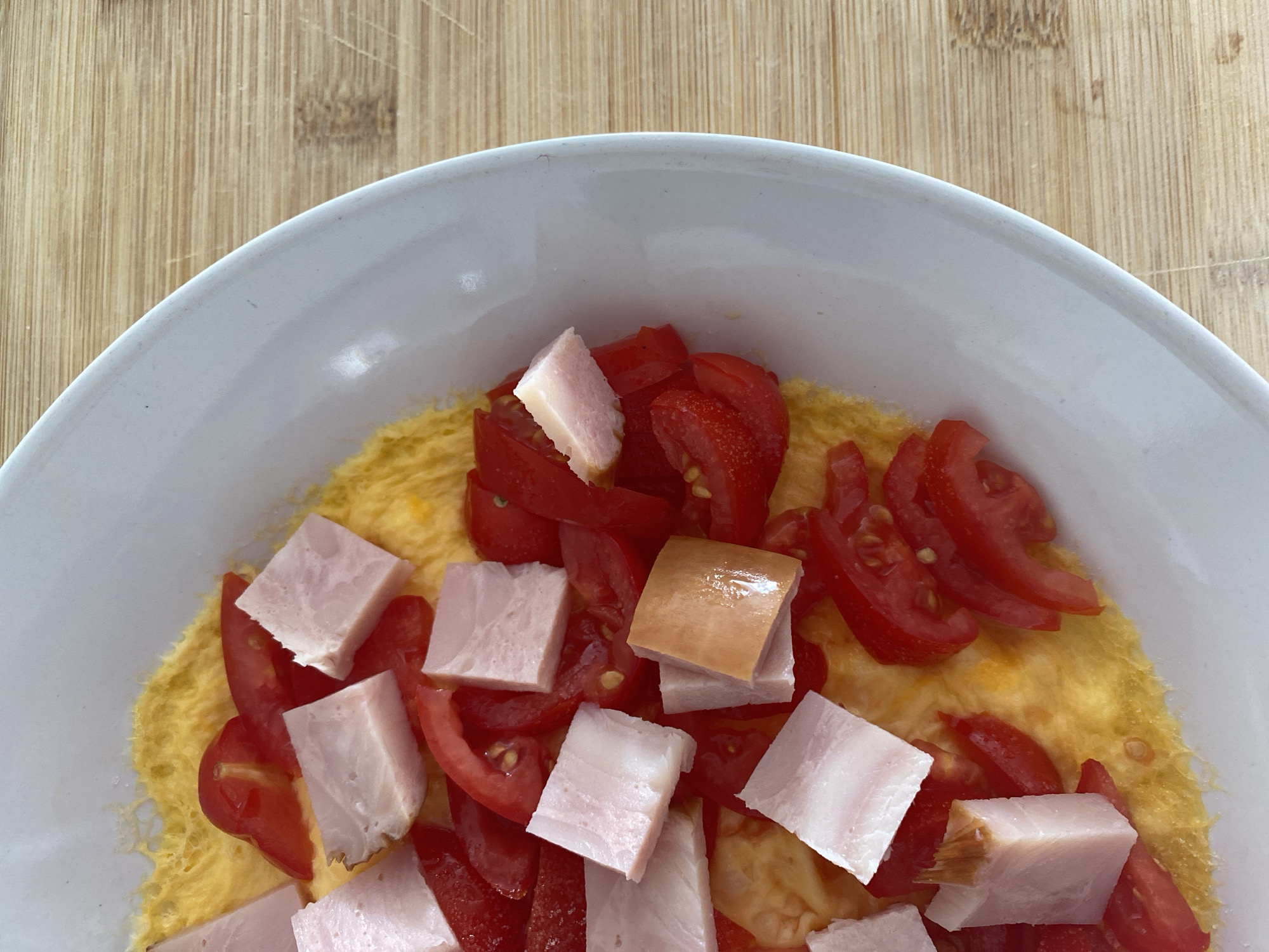 A close-up, overhead shot shows a white plate with an omelet topped with diced tomatoes and cubed ham. The omelet is golden yellow and appears fluffy. The tomatoes are bright red and cut into small wedges. The ham is pale pink with some white fat, cut into small, roughly square pieces. The plate is positioned on a light brown wooden cutting board. The scene is static and well-lit, with no people or discernible urban elements, suggesting a domestic or culinary setting. The image is cropped to focus on the food, with the edge of the plate and cutting board visible on the right side. There are no indications of Yanino-1, Russia.