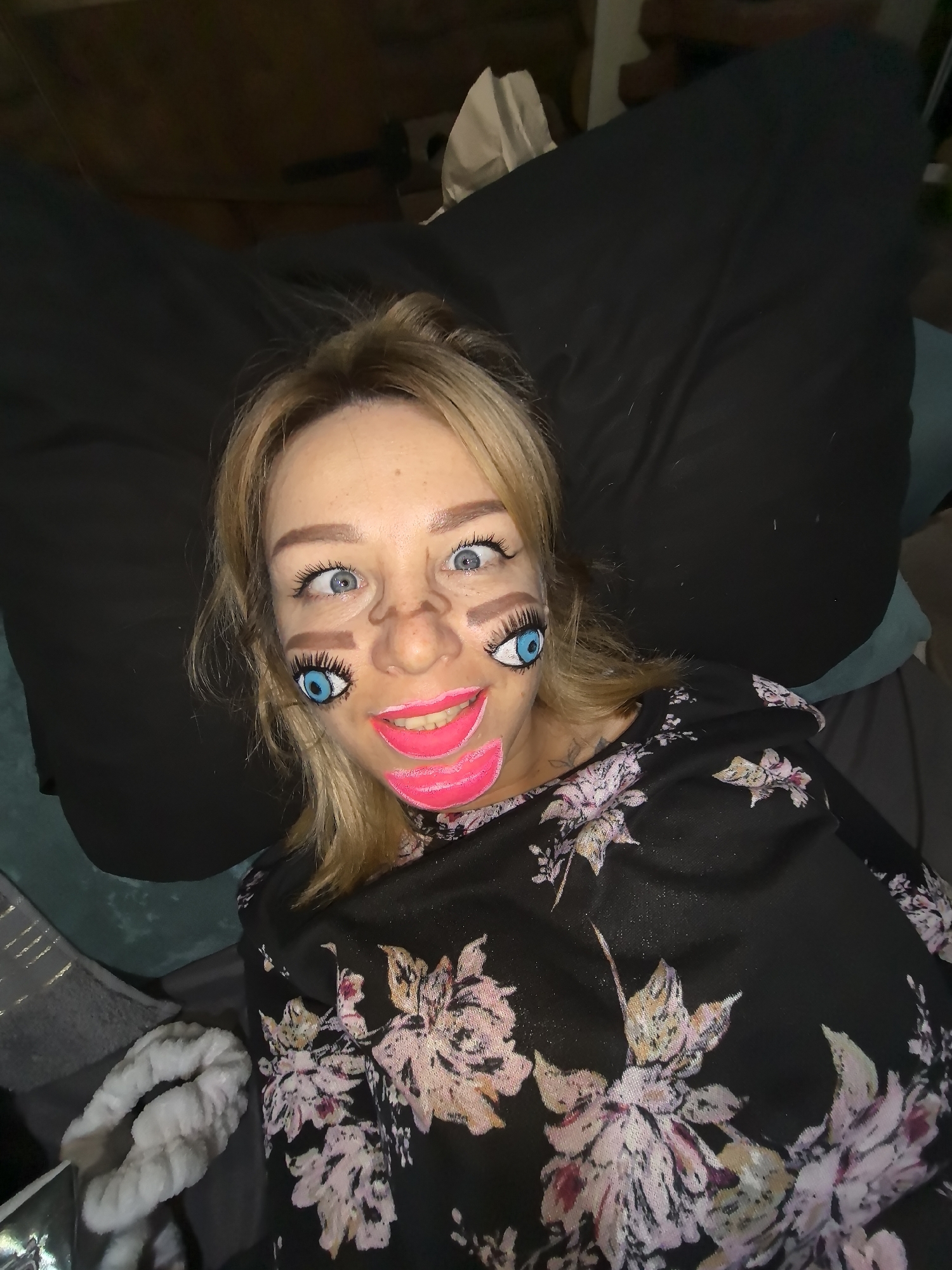 A fair-skinned individual with blonde hair is captured in a close-up, high-angle shot, lying down and looking directly at the camera. The person's face is adorned with elaborate and playful face paint, creating a humorous, cartoon-like appearance. Their actual blue eyes are wide open, appearing slightly crossed or intensely focused.
The face paint features a large, exaggerated brown nose drawn over their real nose. Below each of their real eyes, a vibrant, large blue eye with prominent black eyelashes and white highlights is painted on their cheeks. Above their actual eyes, two small brown shapes suggest additional eyebrows or facial features. Their mouth is covered in bright, hot pink paint, forming a wide, exaggerated smile with visible white teeth. Additional brown lines are painted on their forehead and cheeks, further enhancing the caricature effect.
The person is wearing a black top with a distinctive floral pattern, featuring pink, white, and yellow/gold blossoms. They are positioned against a dark, likely black, pillow or cushion, with another dark blue or teal cushion partially visible to the left. The background is dark and indistinct, suggesting an indoor setting, possibly a bedroom or living room, under dim lighting, likely in the evening or night. In the bottom left corner, a white, fluffy, textured object, possibly a headband, is visible. There is no visible text in the image. The overall impression is one of artistic expression, playfulness, or a costume.