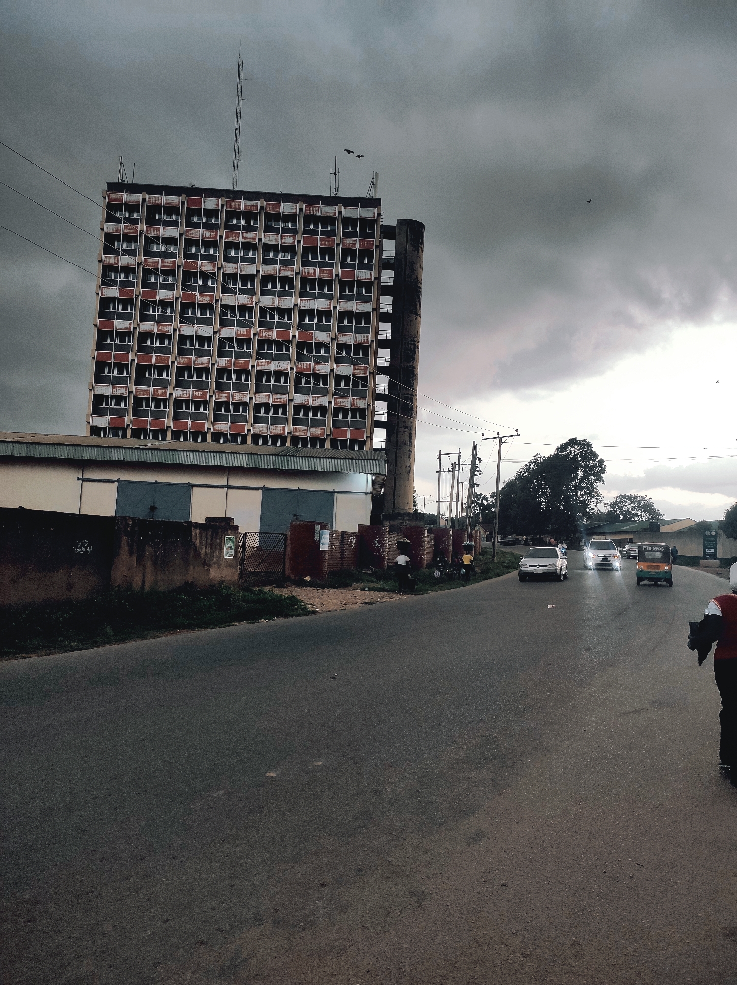 The image depicts a street scene in Jos, Nigeria, under a cloudy sky. A tall, multi-story building with a grid-like facade dominates the left side of the frame. Below it is a lower, one-story structure with a corrugated metal roof, partially obscured by a low wall. The road stretches into the distance, with several vehicles, including cars and a commercial tricycle, visible. A person is partially visible on the right edge of the frame. Power lines stretch across the top of the image.