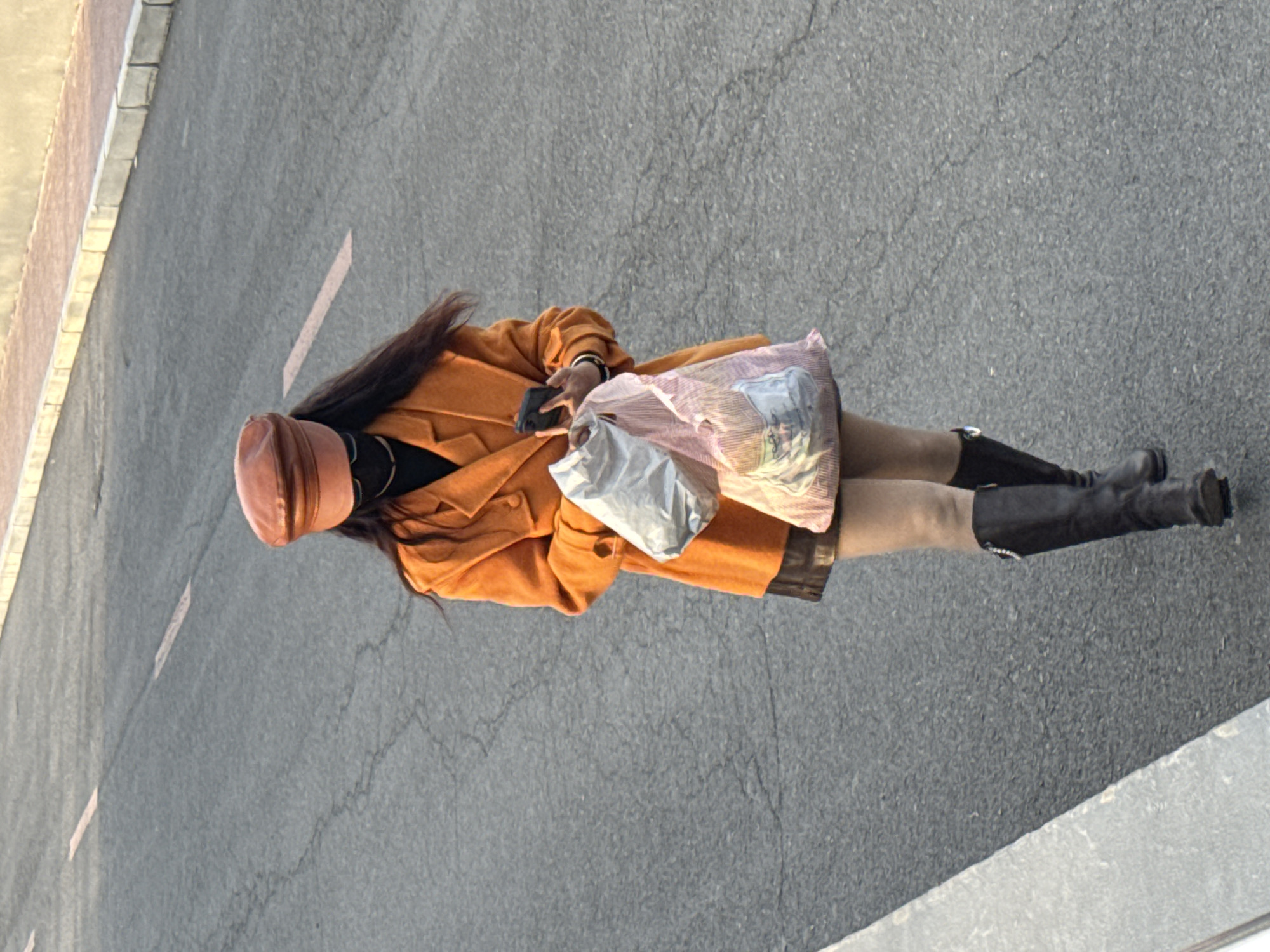 The image, taken from an overhead perspective, depicts a person standing or walking on an asphalt road in Zhongtun, China, under bright daylight conditions. The subject, likely female, has long dark hair and is wearing a warm, autumnal ensemble. Her attire includes an orange or rust-colored long coat over a black top, a short dark skirt, and light-colored opaque tights or leggings. She is wearing black knee-high boots and a reddish-brown flat-top cap.
In her right hand, the person holds a black smartphone. With her left arm, she carries two shopping bags: one is a striped pink and white plastic bag with some indistinct graphics, and the other is a plain white or light grey opaque plastic bag. The background consists of a textured asphalt road, with a faint reddish line painted on its surface behind the person. To her left, a light-colored concrete sidewalk or curb is visible. No specific activity beyond walking or standing is clearly indicated. No discernible text is visible on any of the objects.