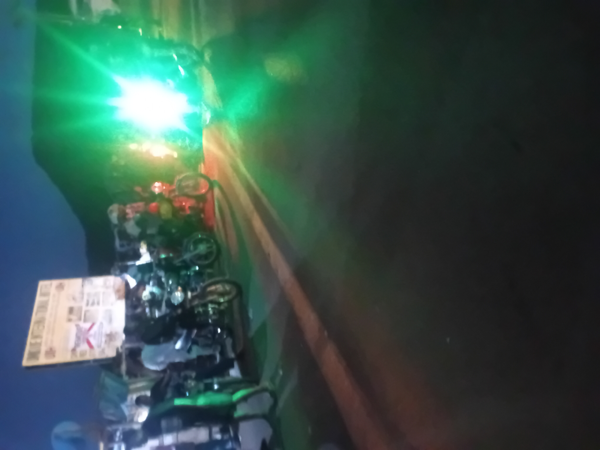 The image, captured at night in Lokoja, Nigeria, is vertically oriented, dark, and blurry, exhibiting significant lens flare from a bright light source.
The scene primarily features several two-wheeled vehicles, likely motorcycles or bicycles, parked or arranged closely together. These vehicles occupy the lower-left and central parts of the frame. While most are dark and indistinct due to the low light and blur, one vehicle appears to have a red body. Components such as handlebars and wheels are faintly visible.
A prominent, intense green light source dominates the upper-left quadrant of the image. This light emits a powerful, almost star-like glare with green rays extending outwards, illuminating the surrounding area with a green tint. This green light casts a strong flare across the image, especially over the vehicles below it.
On the far left, towards the center-left, a light-colored, rectangular sign or poster is visible, appearing to be mounted vertically. It's quite blurry, but a red and white emblem, possibly a flag or logo, can be distinguished in its center. Faint, illegible text and several small images are also present on the sign. The words "MAY CENTENIAL" are barely discernible at the top.
To the right of the vehicles, a dark, reddish-brown surface extends, possibly a wall, fence, or part of a building structure. The ground beneath the vehicles and extending into the lower right is a dark, indistinct surface. No identifiable individuals are visible in the image. The overall setting appears to be an outdoor urban or semi-urban area, likely a street or parking lot, at night.