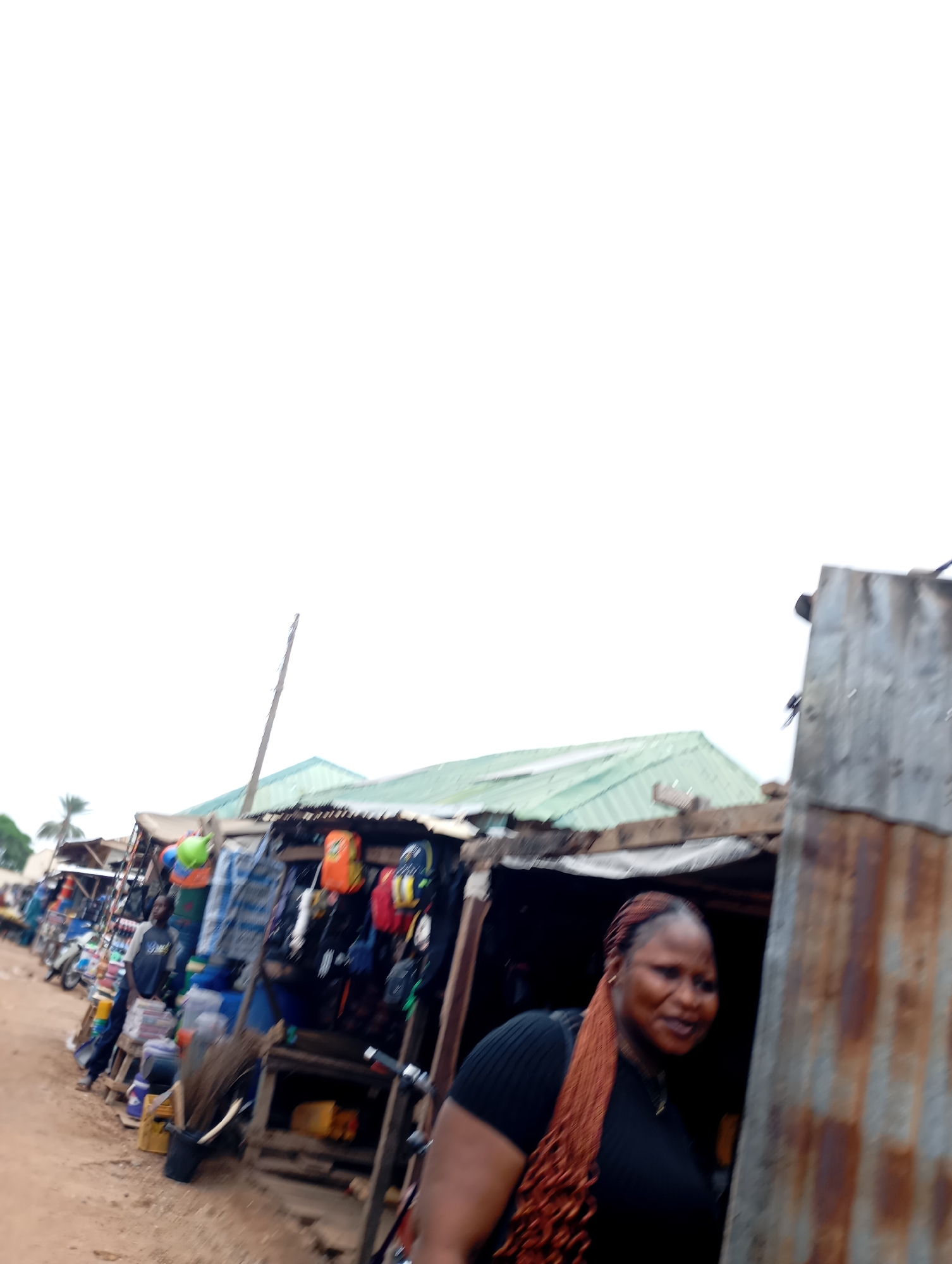 The image shows a street market in Shendam, Nigeria. In the foreground, a woman with long, braided reddish-brown hair is partially visible, wearing a black top. She is standing to the right of the frame, facing left and slightly away from the camera, with a smile on her face. Behind her, a stall made of corrugated metal displays various goods, including bags, clothing, and what appear to be food items. To the left of this stall, a row of other market stalls stretches into the background, with tarpaulin roofs and a variety of merchandise. Motorcycles and other items are visible in the distance, indicating a busy marketplace. The ground is unpaved, suggesting a rural or semi-urban setting. The sky is overcast and bright white.