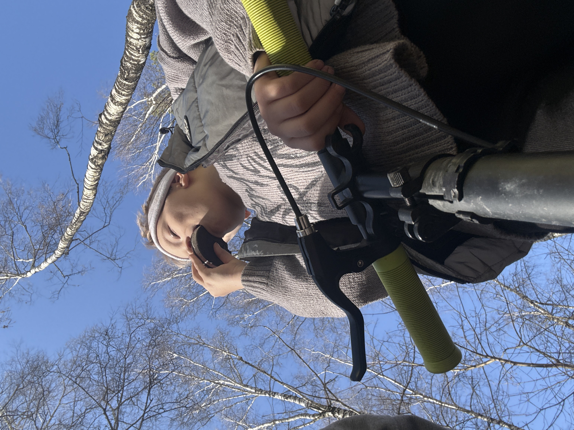 A low-angle shot depicts a person holding a disc-shaped object to their ear, seemingly in conversation. The scene is set outdoors, evidenced by the presence of bare tree branches against a clear blue sky, indicating possibly late autumn or winter. The person is wearing a light gray knitted sweater and a grayish-green vest, and a headband. They are positioned behind the handlebars of a scooter or bicycle, with a lime green grip, a gray metal bar, and a black brake lever visible. The photo's angle suggests it was taken from a ground-level perspective, looking upwards toward the individual and the sky. The specific location details of Dmitrov, Russia are not discernible from the image.