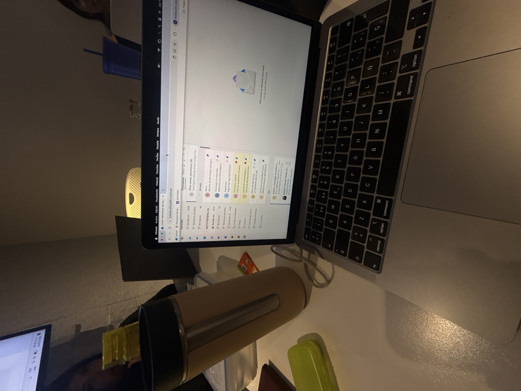 The image shows a desk with a laptop, a thermal mug, and other small containers. The laptop screen displays an email inbox. Email addresses are visible in Spanish, suggesting a Spanish-speaking location. The laptop is an Apple Macbook with a black keyboard. The thermal mug is a light brown color with a dark brown band around the top. The background includes a partial view of a person, a blue plastic cup or container, and a yellow mesh chair. Based on the language displayed on the computer and the details captured, it is likely that the photo was taken somewhere in Mexico, possibly Mexico City.