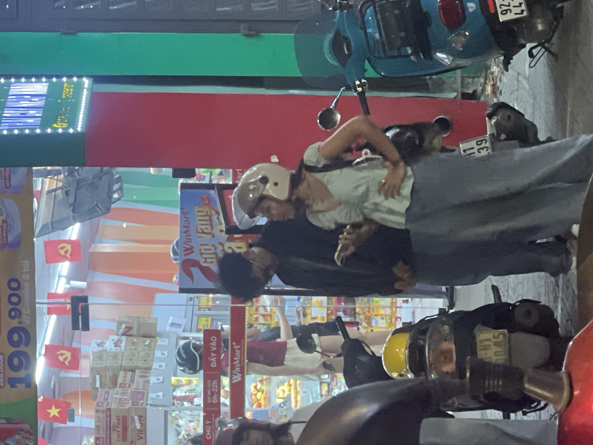 This image depicts a street scene in Da Nang, Vietnam, likely at night, given the artificial lighting. In the foreground, a portion of a red motorcycle is visible, suggesting the viewer is positioned behind it. To the left, a person wearing a white top and a maroon hat is partially visible. Adjacent to them is a black motorcycle with a yellow helmet on its rear.
The central focus of the image is a young couple standing near the entrance of a WinMart convenience store. The woman, wearing a light green V-neck top and grey pants, has her hands on her hips and is looking to her left. She is wearing a light-colored helmet. The man beside her, dressed in a black jacket and dark pants, is looking down at his phone and appears to be holding it in his right hand.
Behind them, the entrance of the WinMart store is visible. The store displays various products, including boxes of food items and what appear to be bottles of oil or beverages. Red and yellow flags, some featuring a yellow star, are hung above the store entrance. A sign above the store reads "WinMart" followed by "Giờ Vàng" (Golden Hour). Further to the right, a bright green and red facade of a building is visible, along with a blue scooter. A digital billboard with blue and yellow lettering is mounted on the green section of the building. The ground is paved.