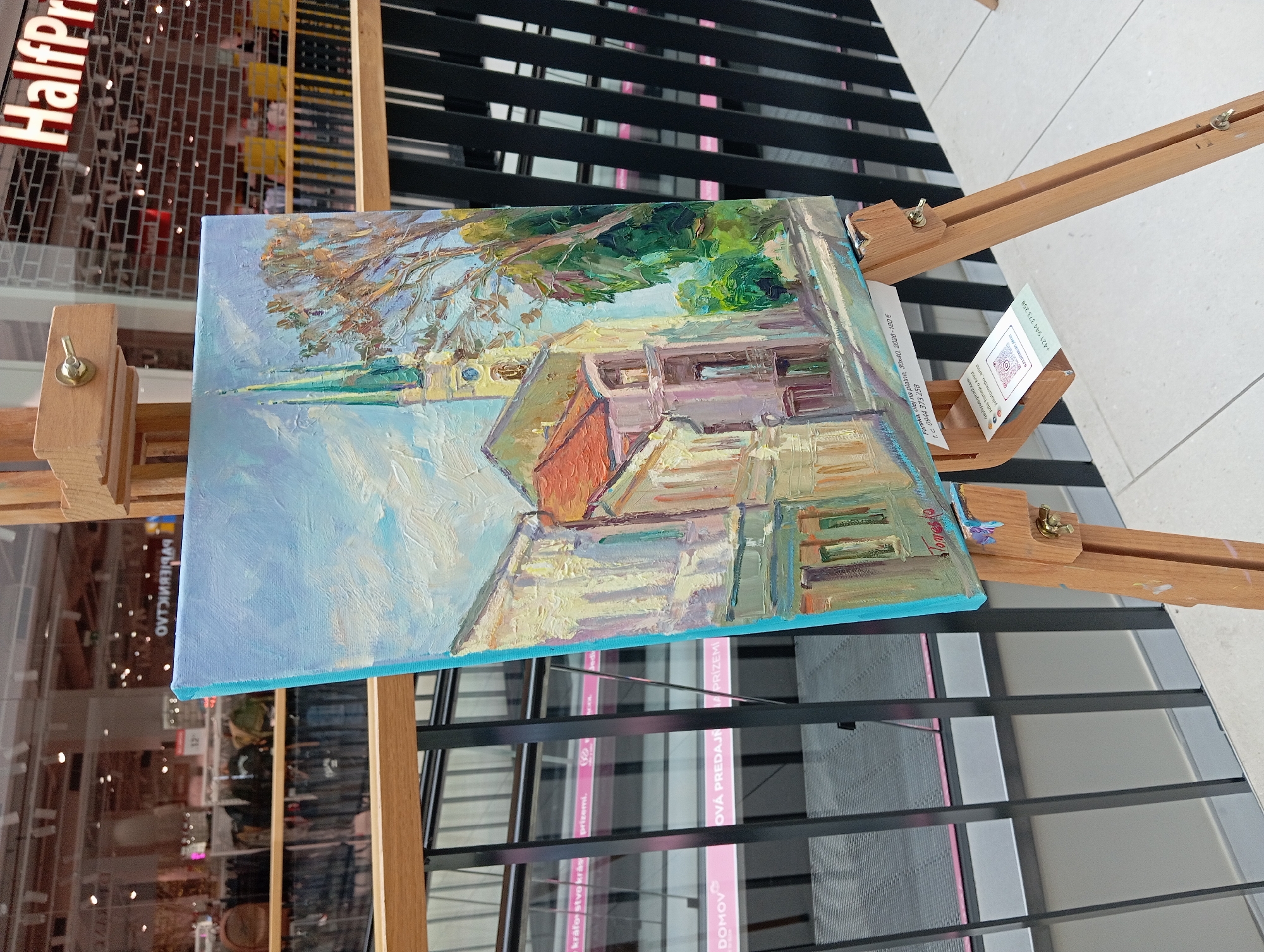 A painting of a European street scene featuring buildings with red tiled roofs and a church tower with a green spire is displayed on a wooden easel. The painting is positioned in front of a large glass window that looks into a shopping mall. The mall interior is visible, with rows of clothing racks and a sign that reads "HalfPrice" in large red letters. The painting's signature appears to be "Tomaso". A label attached to the easel provides details about the artwork, including its title "Farske, olej na plátne", dimensions "30x40", year "2023", and price "180 €", along with a phone number and website. Another small card displays a QR code and social media handles. The overall environment suggests a public space, likely within the shopping mall. The painting is the central subject, with the mall serving as the backdrop.