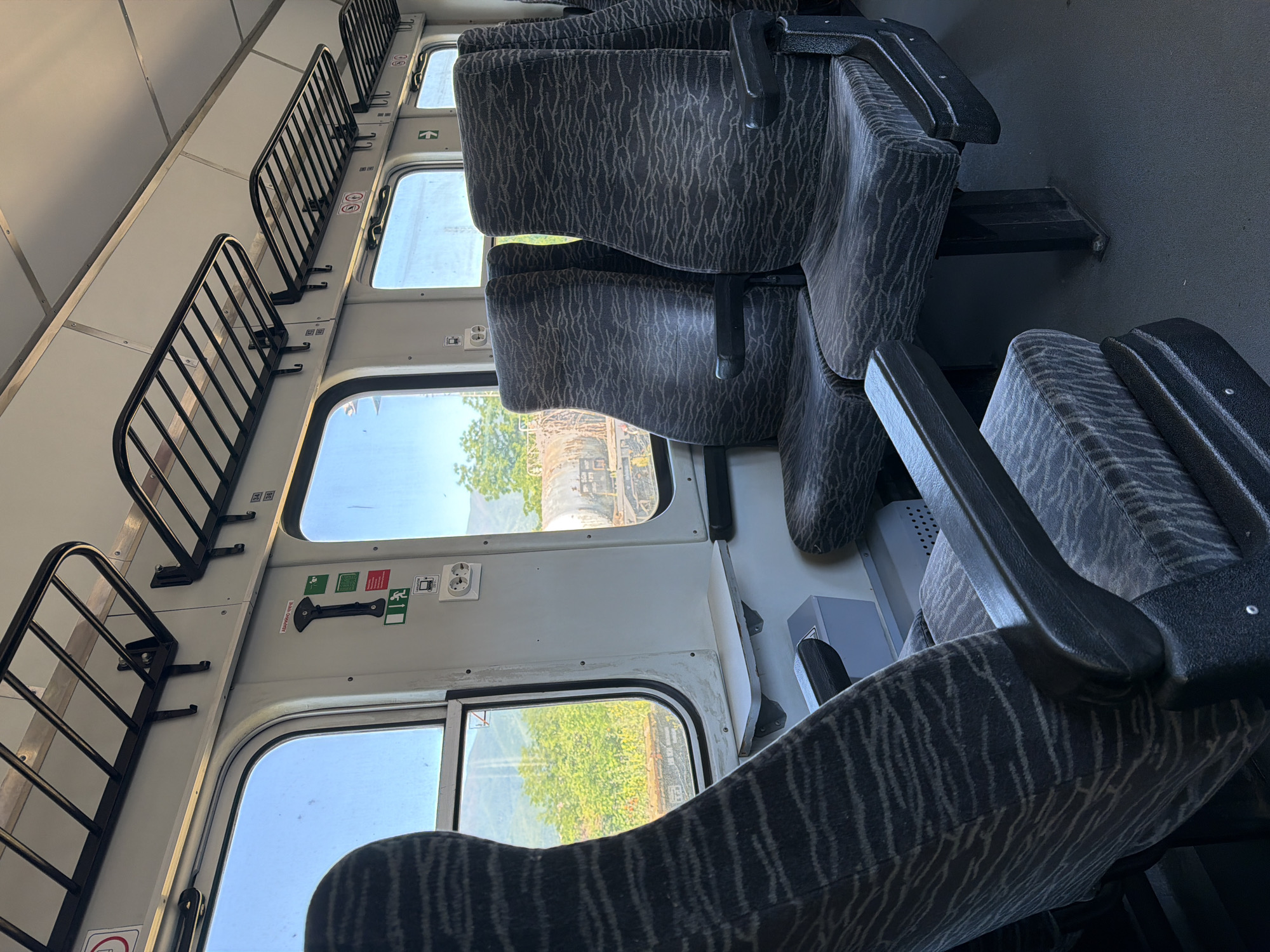 The image shows the interior of a passenger train car. Several rows of upholstered seats with armrests are visible, and overhead luggage racks line the wall above the windows. Outside the windows, trees and a smokestack can be seen, suggesting an industrial area. The location is near Septemvri, Bulgaria, as determined by the smokestack viewed from the train car. No people are visible in the image.