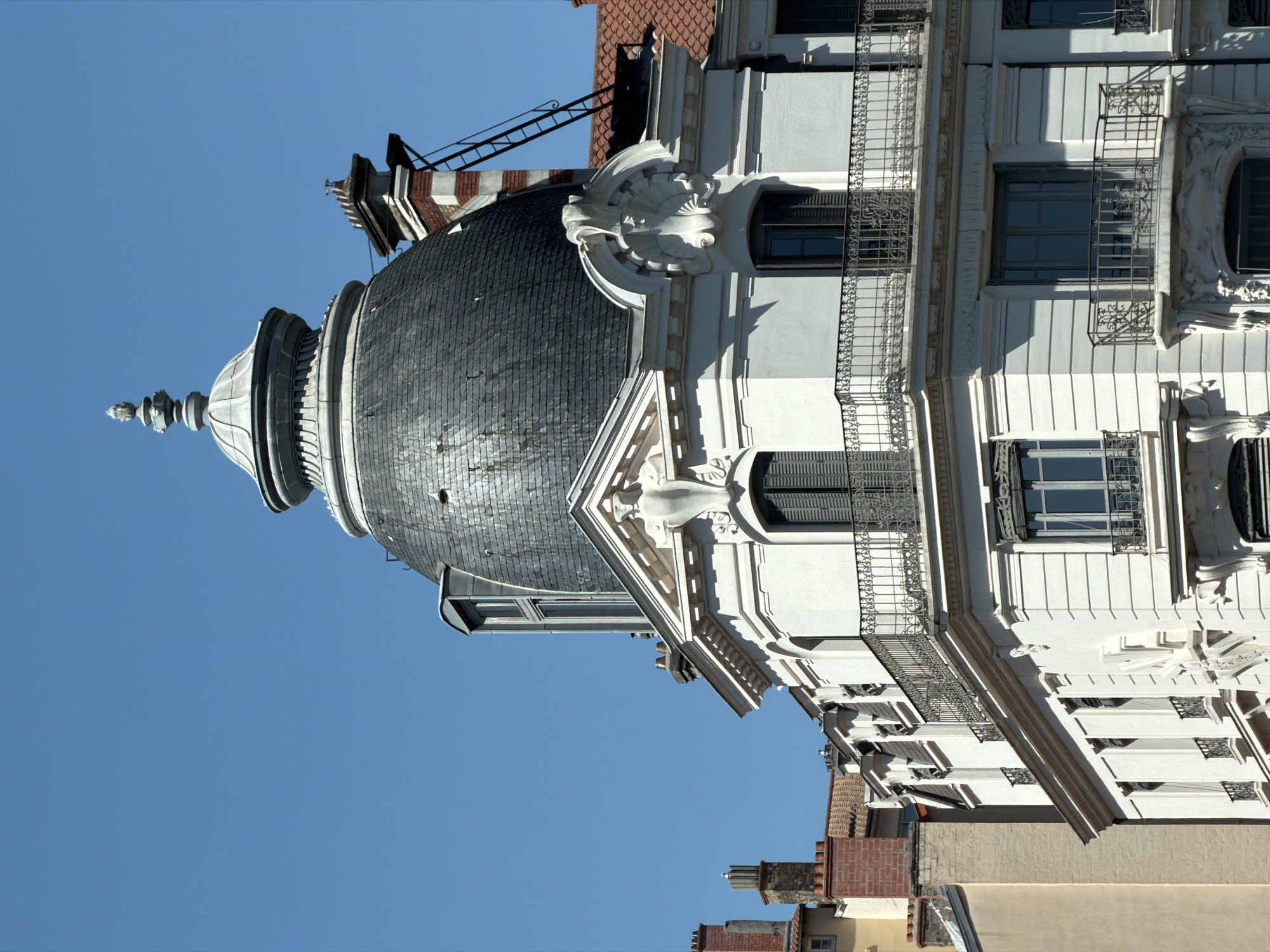 The image shows a close-up view of the top floors of a white, ornate building with a dark, domed roof, characteristic of Belle Époque architecture. The building is located in Lyon, France. The dome is covered in slate tiles and topped with a decorative finial. Below the dome, the building features multiple stories with balconies adorned with intricate wrought-iron railings and classical decorative elements around the windows. The windows are tall and narrow with dark shutters. In the background, a clear blue sky dominates, with glimpses of other buildings and chimneys suggesting an urban setting.