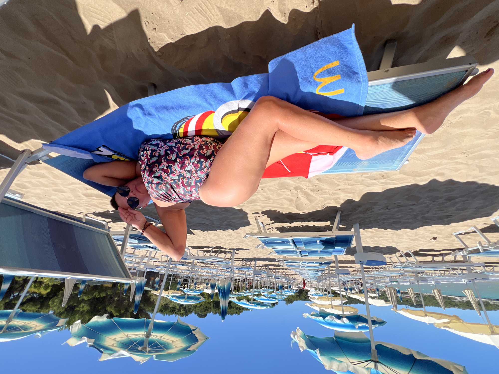A woman in a patterned swimsuit lies on a blue lounge chair on a sandy beach. She is positioned on her side, with her legs extended and crossed at the ankles. The lounge chair is covered with a blue towel that has a yellow "M" logo on it and a colorful, circular design. In the background, rows of empty lounge chairs and blue and white striped umbrellas are set up on the beach. The sea is visible in the distance, and a line of trees borders the top of the beach. The sky is clear and blue. The scene appears to be located on a beach in Peschici, Italy, a popular tourist destination known for its beautiful beaches and coves.