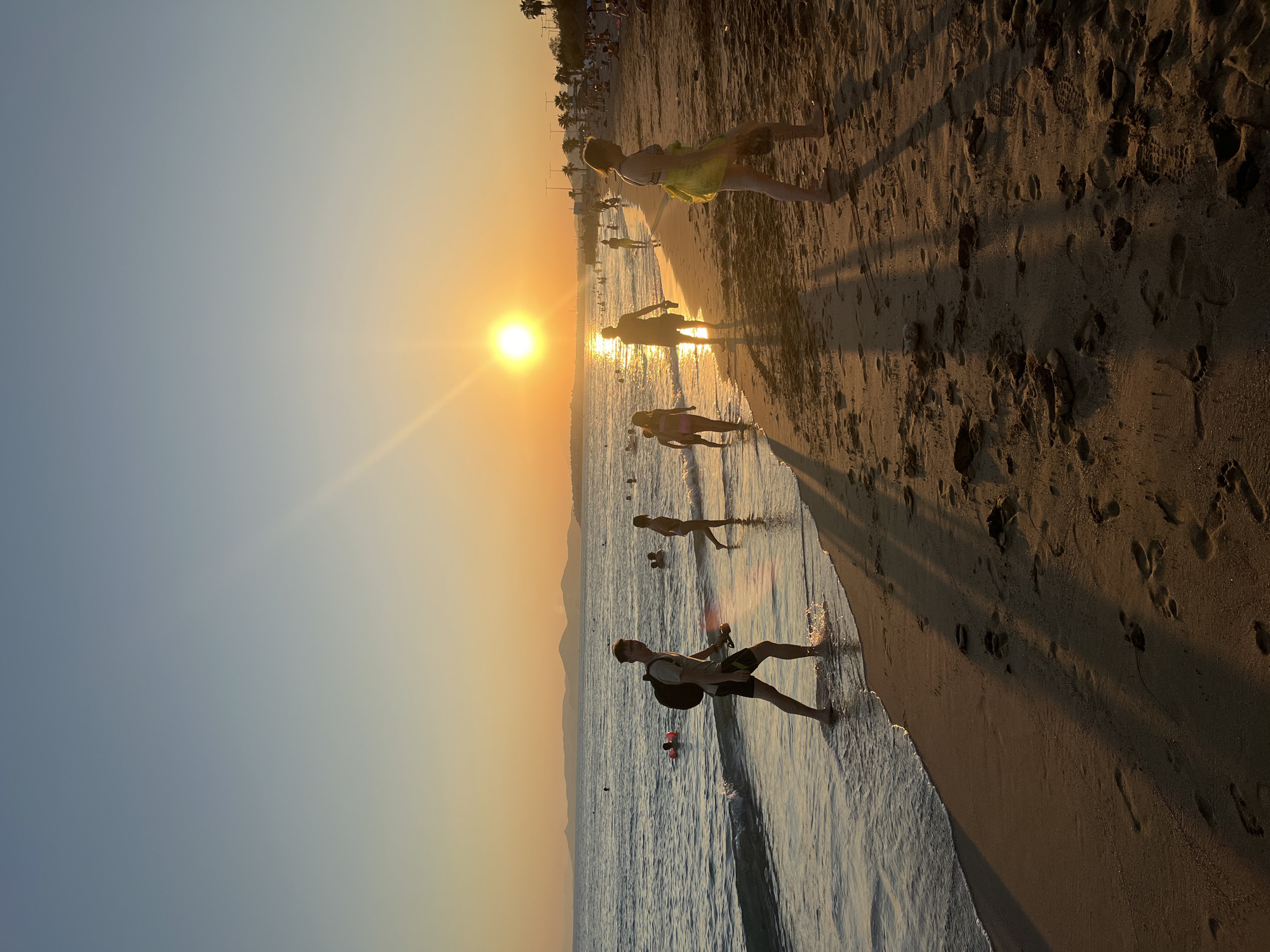 This is a photograph of a beach at sunset, likely in Paleo Faliro, Greece, given the distant hills and the clear sky. The sun is low on the horizon, casting a warm, golden light across the scene and creating long shadows on the sand. Several people are visible on the beach and in the shallow water. In the foreground, a young man walks along the water's edge, his wet shorts and bare feet indicating he has been in the sea. He carries a backpack and a yellow object. Further down the beach, other figures stroll and play in the water. The sea is calm with gentle waves lapping at the shore. The sand is scattered with footprints. In the background, the coastline is visible with hills and palm trees, suggesting a Mediterranean setting. The overall atmosphere is one of leisure and relaxation during a warm evening.