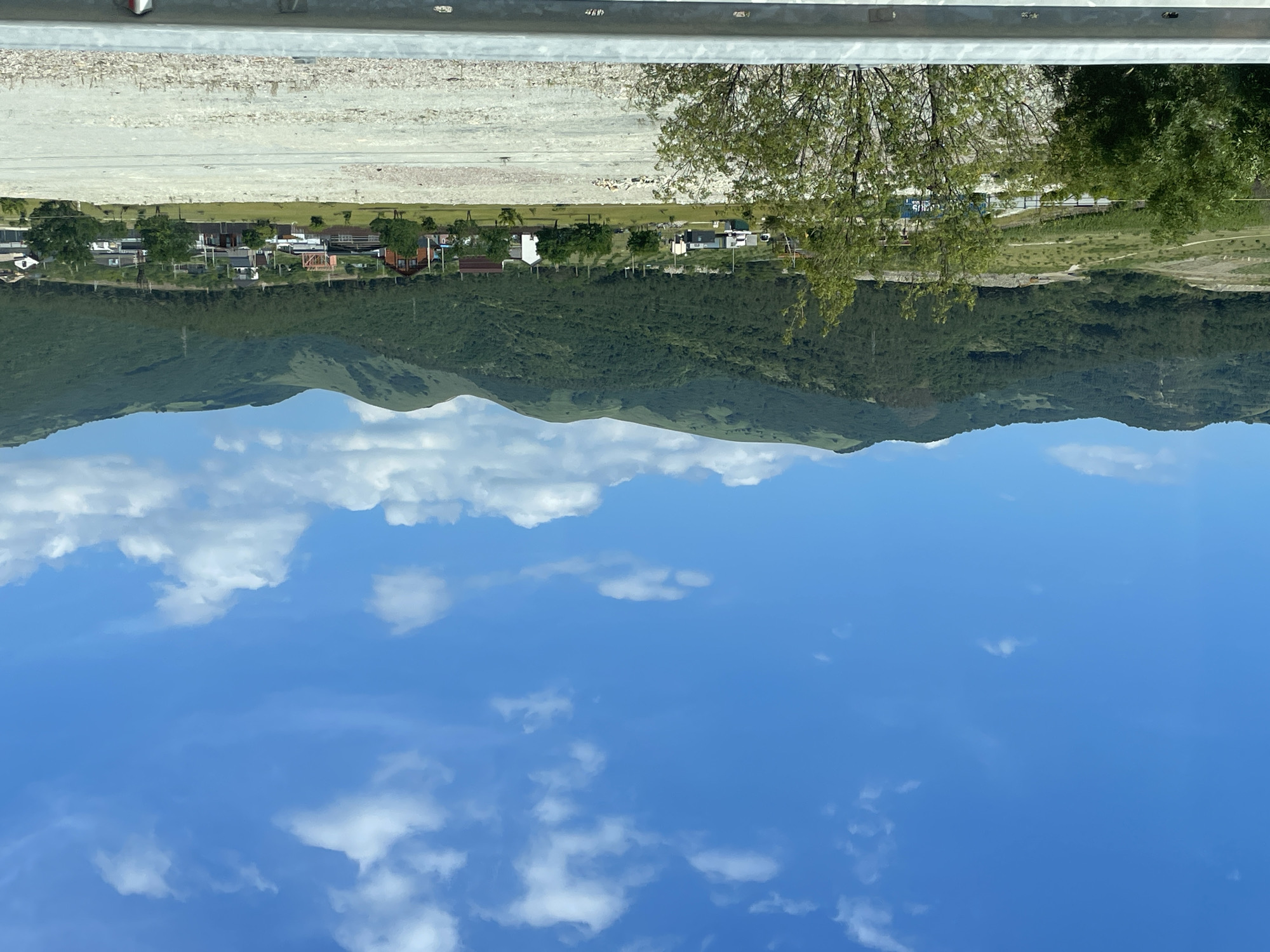 This is a vertical, wide-angle, eye-level shot of a residential area at the foot of a mountain range, under a bright blue sky with scattered white clouds. The image is oriented such that the sky appears at the bottom and the land at the top.
In the foreground, at the bottom of the image, is a vast expanse of blue sky, dotted with fluffy white clouds.
The middle ground features a densely forested mountain slope, with varying shades of green indicating the foliage. The terrain appears rugged and undulating. Below the mountain, a line of houses forming a village or small town is visible. The houses are varied in style and color, with some featuring red or brown roofs and others white walls. Trees are interspersed throughout the settlement.
The background, at the top of the image, shows a road or pathway, bordered by a light-colored, gravelly area. A metal railing runs along the top edge of the image, suggesting the vantage point might be from a road or elevated walkway. A tree with green foliage is prominent in the upper right portion of the image.
The scene appears to be a tranquil daytime setting. The bright sky and clear visibility suggest fair weather. The presence of houses and trees indicates a rural or semi-rural residential area. There is no visible text in the image.