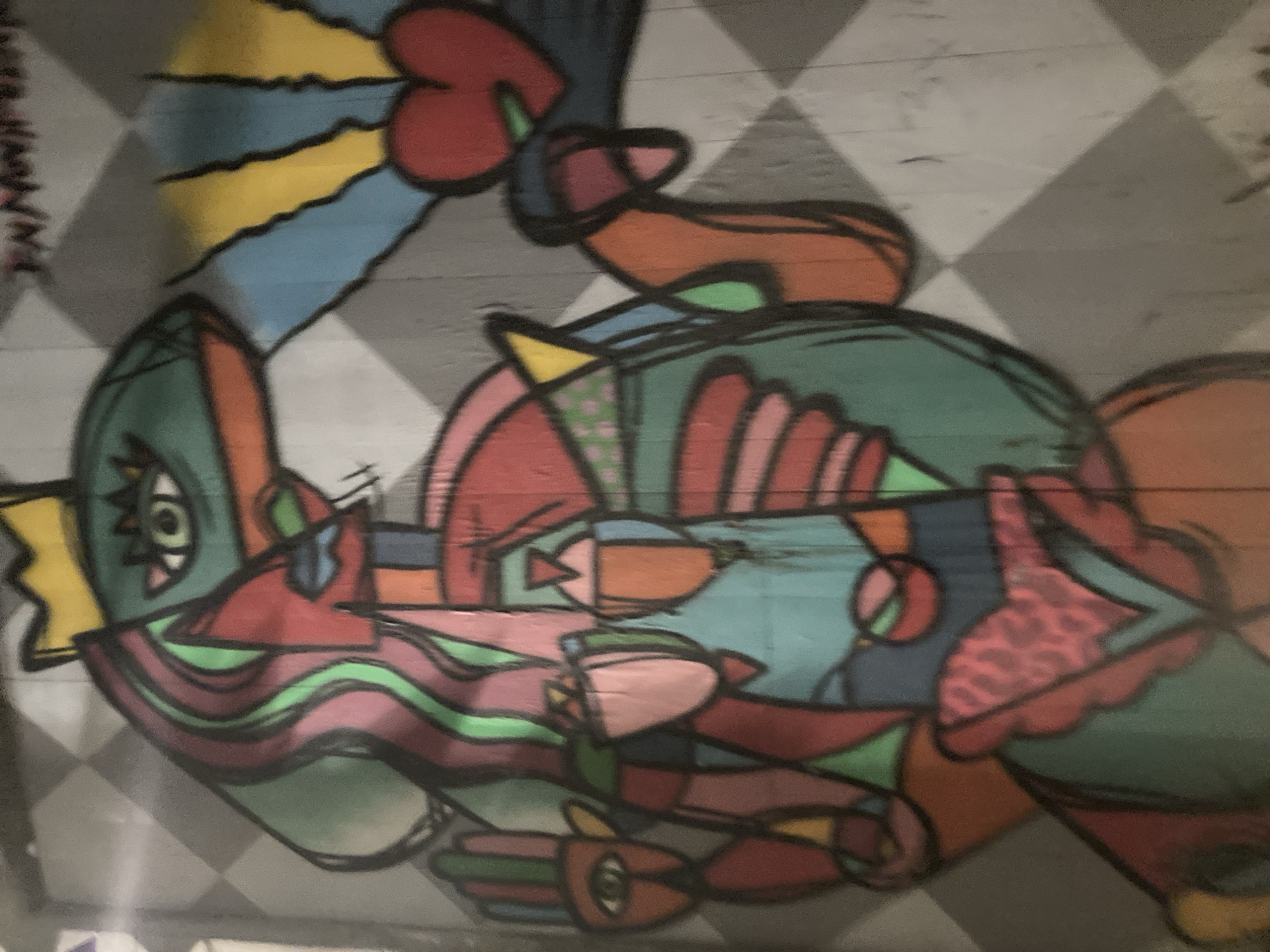 The image shows a colorful, abstract mural on a grey, diamond-patterned wall. The mural depicts a figure with a green head adorned with a yellow crown and a large eye. The figure has flowing, multi-colored hair and appears to be wearing a red and green outfit. One hand is raised, with fingers spread, and features an eye in the palm. The figure holds a red heart radiating yellow light in its other hand. The mural is located in Barcelona, Spain, on a wall near a street. The overall style is bold and geometric, with thick black outlines defining the shapes.