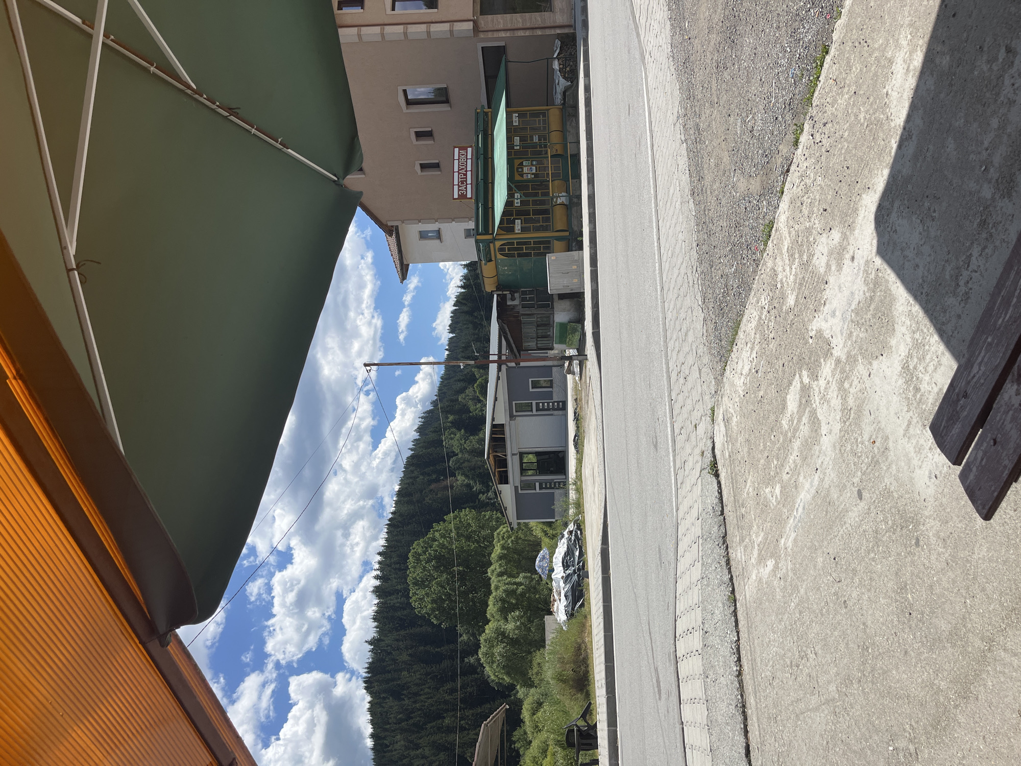 The image captures a street view in Barutin, Bulgaria. In the foreground, a concrete sidewalk is visible, partially shaded by an overhead umbrella structure. Beyond the sidewalk is a road, with a paved surface. On the opposite side of the road, there are several buildings. One building on the right appears to be a two-story structure with windows. Adjacent to it is a smaller, yellow and green building with large windows and a green awning, displaying a sign with Cyrillic text that reads "ЗАСТРАХОВКИ". To the left of this building is a grey, industrial-looking structure. In front of the grey building, there are stacks of covered materials, possibly construction supplies, and a blue and white patterned umbrella. Further in the background, a dense forest covers a hillside under a partly cloudy sky. The scene depicts a rural or semi-rural setting, likely during daylight. There are no people visible in the image.