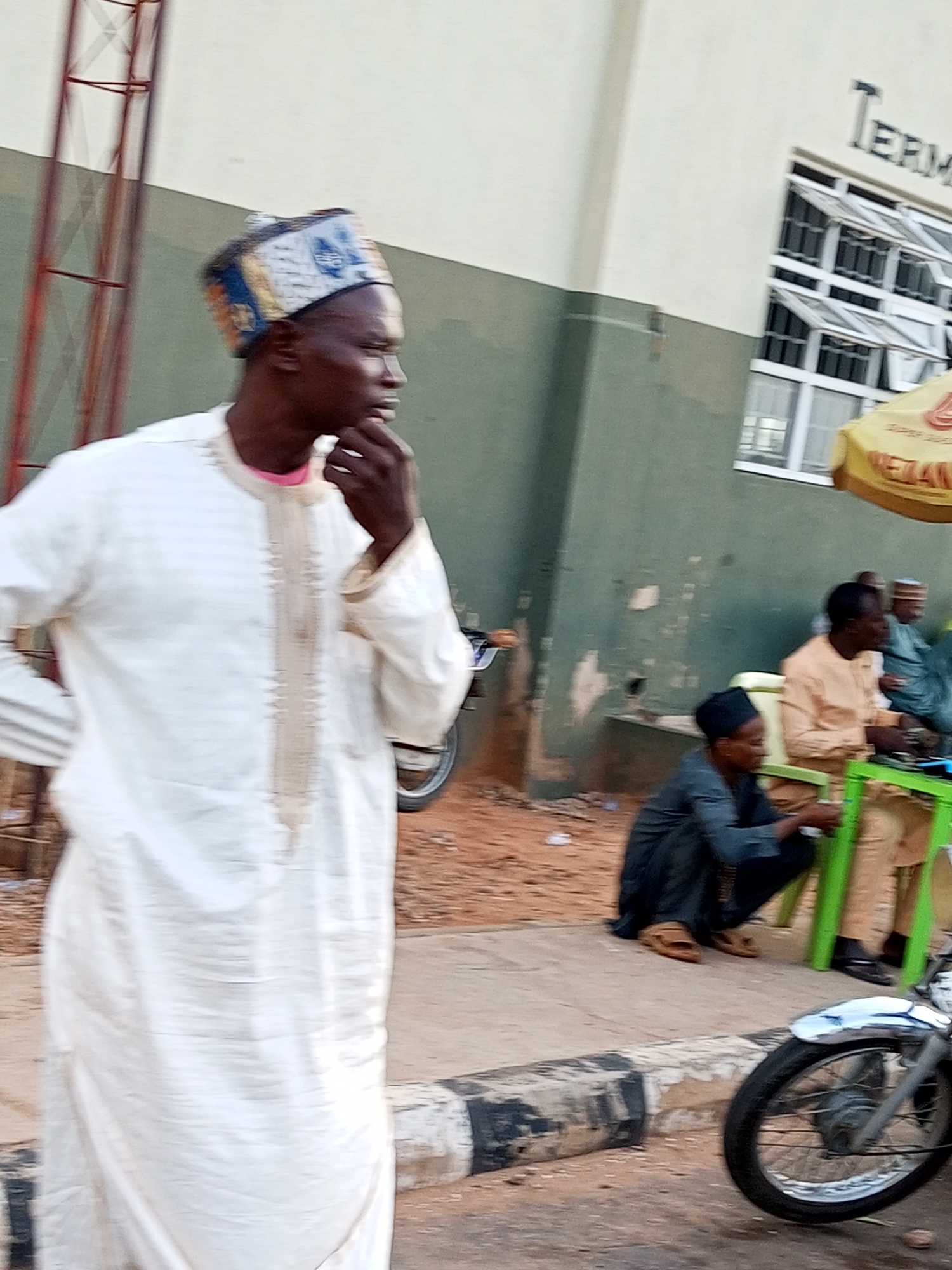 The image depicts an outdoor scene in the city of Gombe, Nigeria. In the foreground, a man stands facing right, wearing a white traditional robe and a patterned cap. His right hand is raised to his chin, suggesting contemplation or conversation. Behind him, other individuals are visible, some seated and others standing near a building with windows and a sign that reads "TERMINAL." A motorcycle is parked on the right side of the frame. The ground appears to be a dirt or paved area, with some debris present. The overall atmosphere suggests a public gathering or waiting area, possibly a bus or taxi terminal.