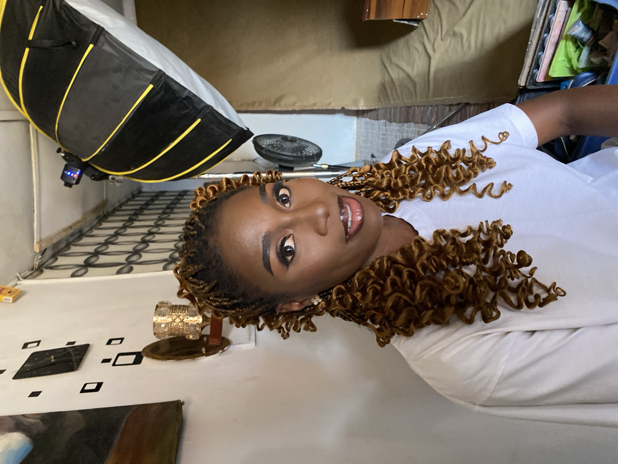 A Black woman is featured in a close-up, chest-up portrait, looking directly at the camera with a confident, slightly smiling expression. She has curly, light brown/auburn hair, styled in what appears to be braids or extensions, and wears a plain white t-shirt. Her makeup includes well-defined eyebrows, eyeliner, and glossy lips. Small, sparkling earrings are visible on her ears.
The setting appears to be an indoor space, possibly a photography studio or a room arranged for a photoshoot. Dominating the left side of the frame is a large, black and yellow softbox or octabox, with a flash unit attached, indicating professional lighting. A smaller, dark, circular object, possibly a fan or additional lighting equipment, is visible behind the woman's head. The floor features a black and white geometric pattern on one side and a plain white surface on the other. In the background, there is a dark brown wooden piece of furniture and a light brown fabric surface, which could be a curtain or backdrop. A small, golden decorative item with intricate detailing is visible on a surface near the patterned floor. A small yellow box is also on the floor.
The woman is actively posing for the photograph, illuminated by the artificial lighting setup. No legible text is visible in the image.