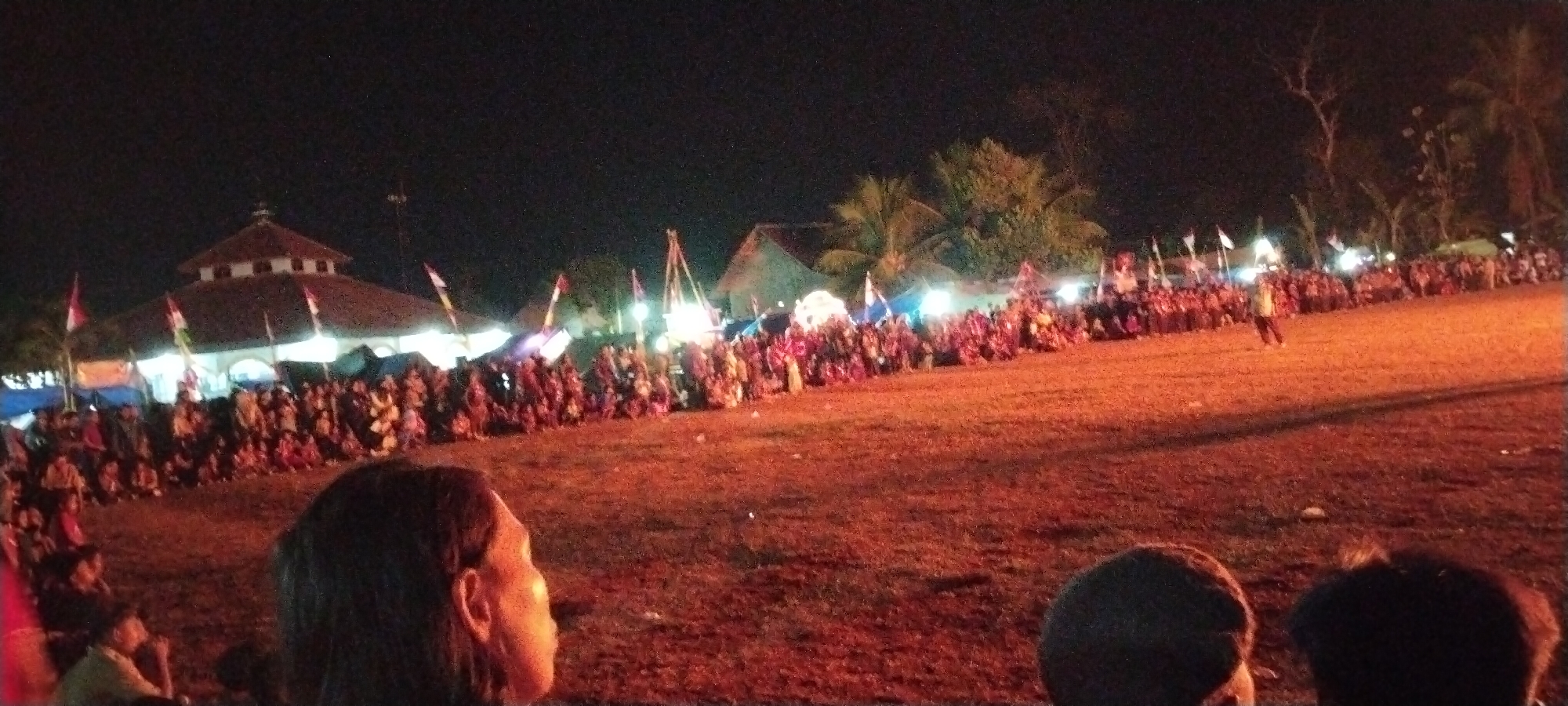 The image depicts a nighttime outdoor gathering in Sido Rahayu, Indonesia, likely a festival or event. In the background, a large building with a domed roof, possibly a mosque or community hall, is illuminated. Rows of people are seated or standing in front of the building, facing a central area. Flags, including red and white ones, are visible among the crowd and structures. The ground appears to be unpaved and covered with dry grass or dirt, illuminated by warm lighting that casts long shadows. Several figures are scattered throughout the scene, some appearing to be performers or attendees. The overall atmosphere is lively and communal.
