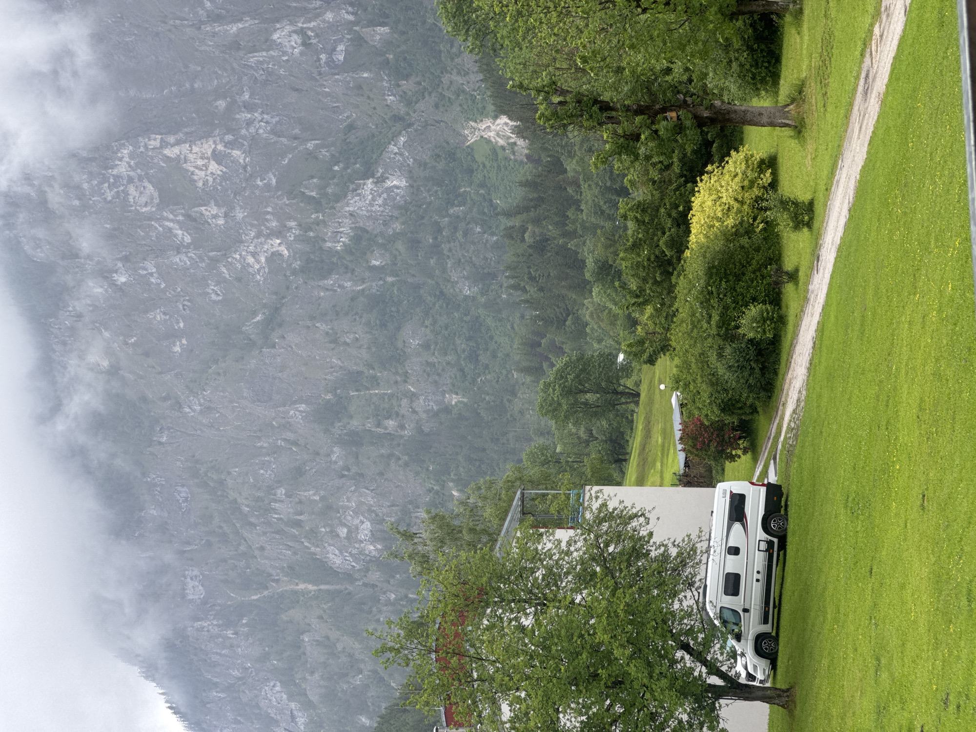 The image, originally rotated 90 degrees clockwise, displays a serene mountainous landscape near Eisenerz, Austria, under an overcast sky.
In the background, imposing, rugged mountains rise, characterized by their grey rocky surfaces and patches of dense green forest. Significant portions of the mountains, especially on the left, are enveloped in mist or clouds, creating a hazy, atmospheric effect and diffusing the daylight.
The middle ground features a variety of trees and shrubs, including a prominent bright yellow-green bush. A light-colored dirt or gravel path gracefully curves through a vibrant green lawn, leading further into the scene. Partially visible on the left is a segment of a white building with a flat roof.
The foreground is dominated by a lush, well-maintained green lawn. Parked on this grass, nestled near the white building and under the canopy of a tree, is a modern white camper van. The word "GLOBECAR" is faintly visible on its side, complemented by subtle red stripe detailing and black wheels with silver accents.
The scene portrays a quiet, static daytime moment, likely at a campsite or a property, with no people visible. The weather appears cloudy or misty, contributing to a tranquil and natural ambiance. The overall impression is one of peaceful immersion in nature.