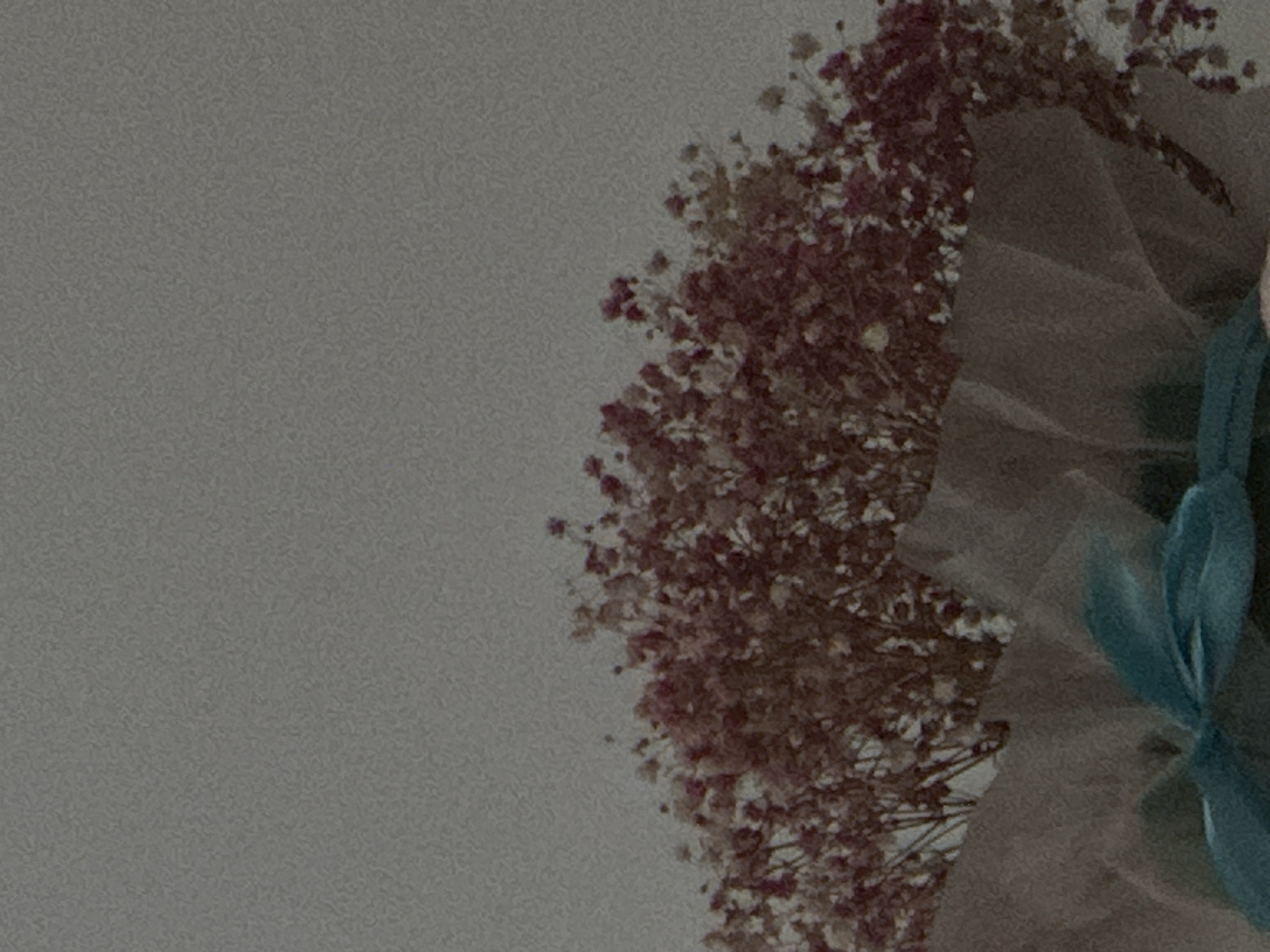 A bouquet of dried baby's breath flowers, with pink and white blooms, is presented in soft, layered brown and white paper wrapping. A teal ribbon is tied in a bow at the front of the bouquet. The background is a muted, textured gray. The image was taken in Georgiyevsk, Russia.